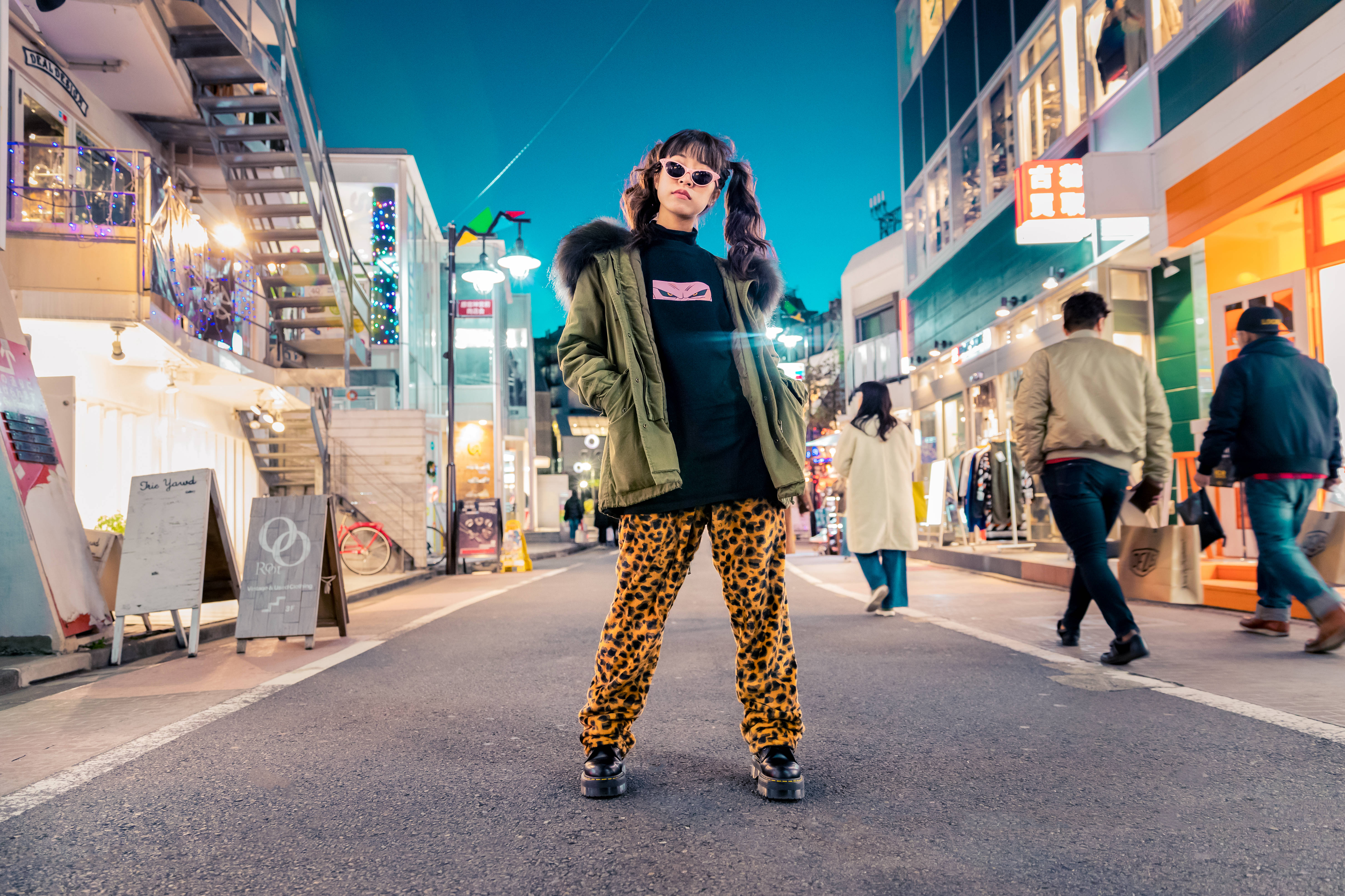 Trendy 桌布 壁紙 — 一位穿著豹紋褲和綠色外套的時尚人士，在黃昏時霓虹燈閃爍的城市街道上自信地站立。