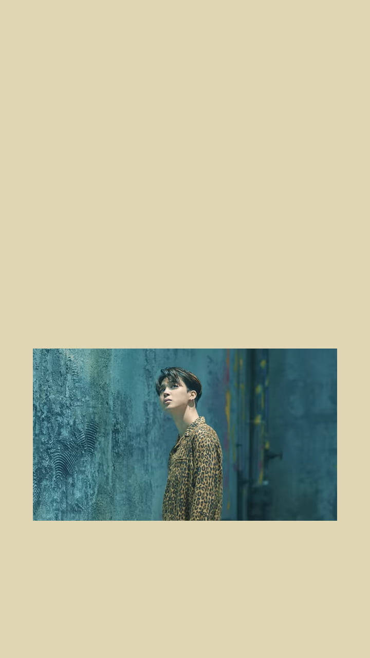 BTS 桌布 壁紙 — 一位穿著豹紋襯衫的年輕男子在柔和日光下靠著帶紋理的藍色牆壁站立。