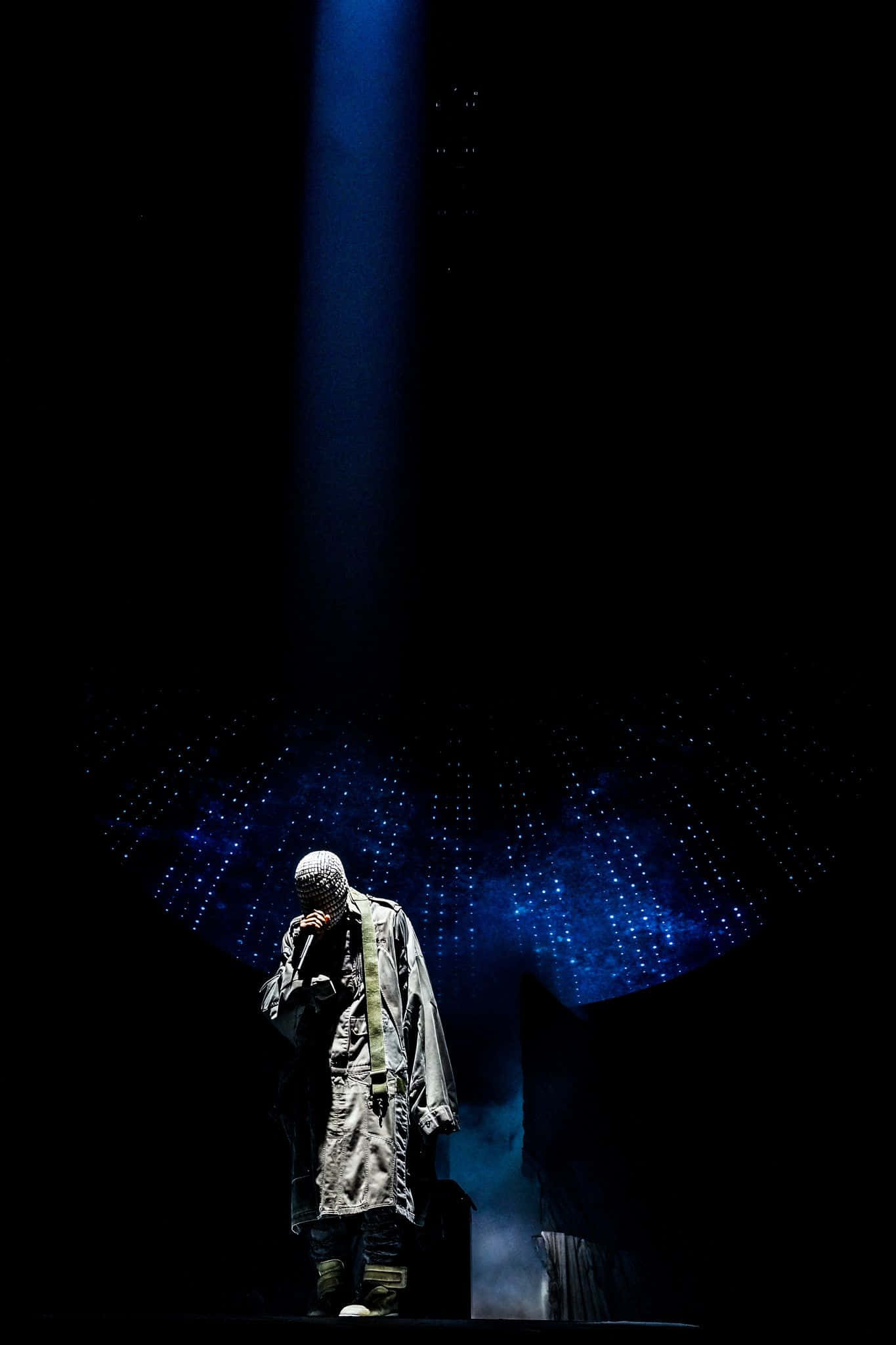 Kanye West Yeezus 桌布 壁紙 — 一位穿著質感外套的表演者獨自站在黑暗的舞台上，沐浴在單一藍色聚光燈下，下方有星狀燈光。