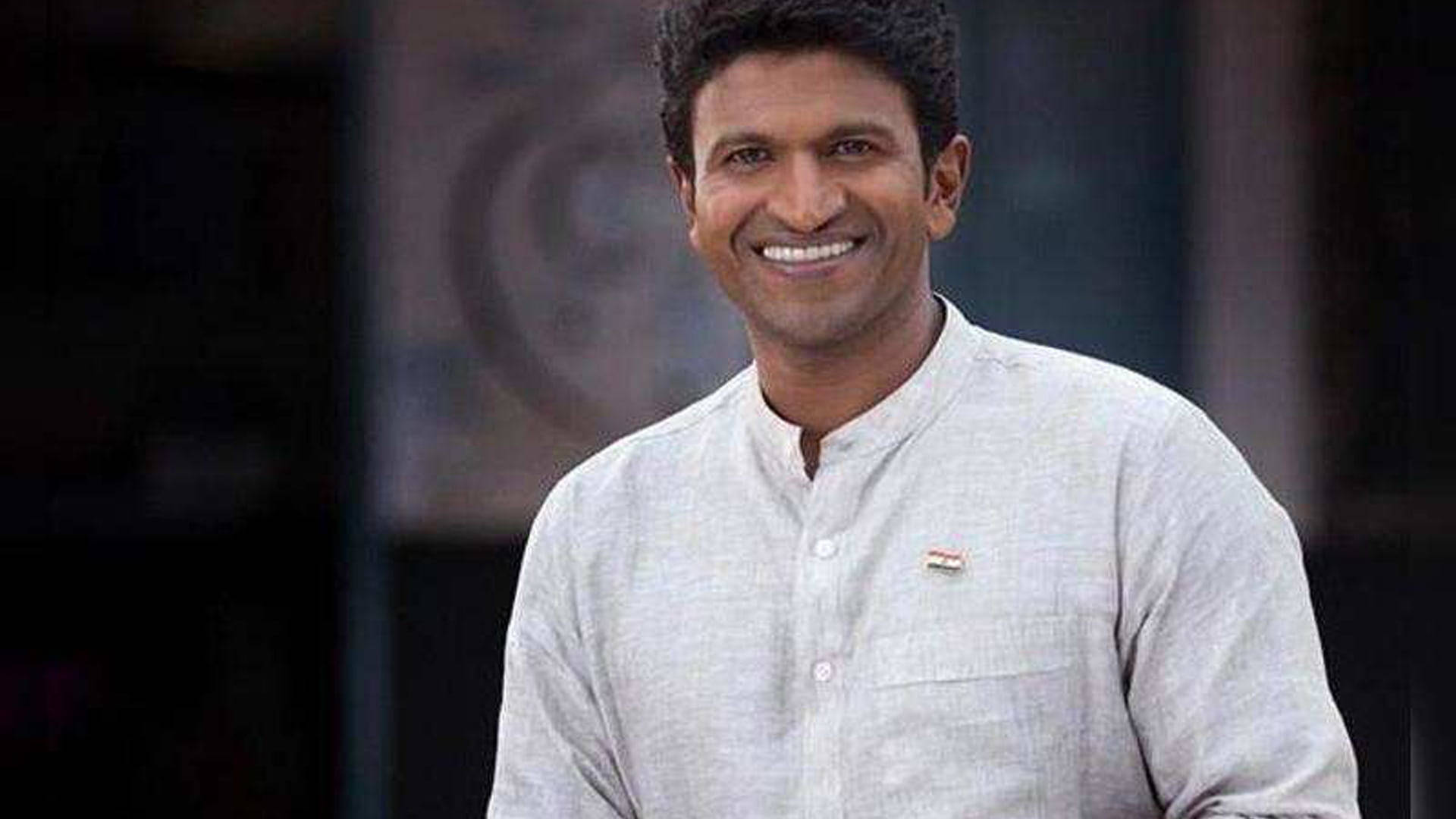 Puneeth Rajkumar 桌布 壁紙 — 一位穿著輕薄亞麻襯衫的開朗男子，在模糊的城市背景下對著鏡頭溫暖地微笑。
