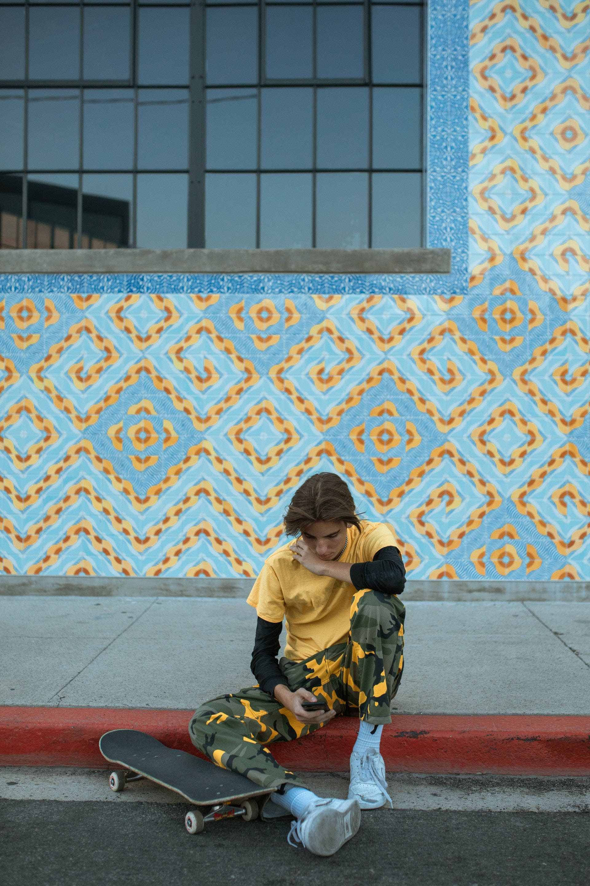 Skater Boy 桌布 壁紙 — 一位穿著迷彩褲和黃色襯衫的年輕人坐在路邊石上，旁邊有一塊滑板，靠著充滿活力的藍色和橙色瓷磚牆，上方是玻璃建築。