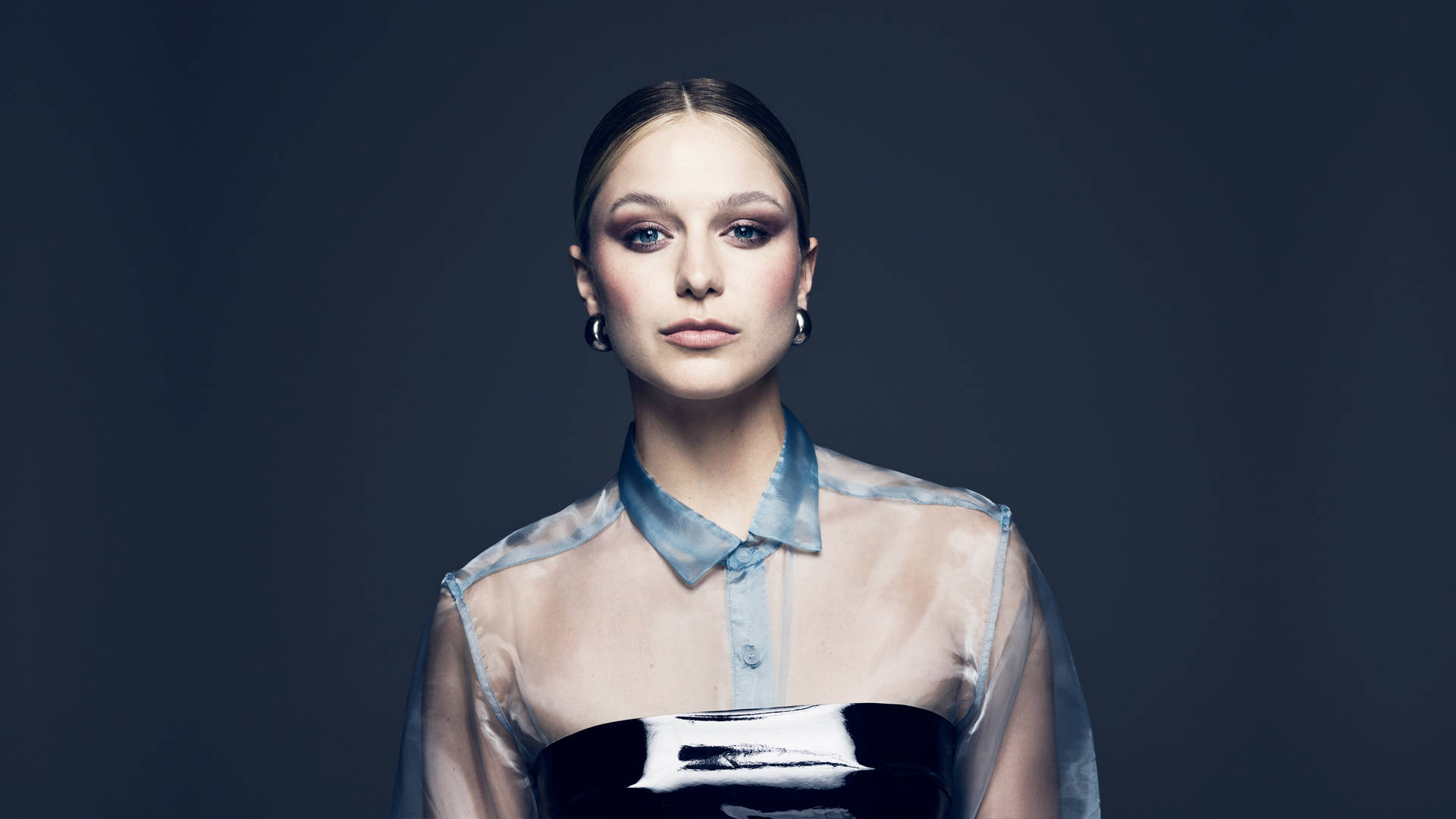 Melissa 桌布 壁紙 — 一位穿著透視藍色襯衫和黑色上衣的優雅女性，在深灰色背景下直視鏡頭。