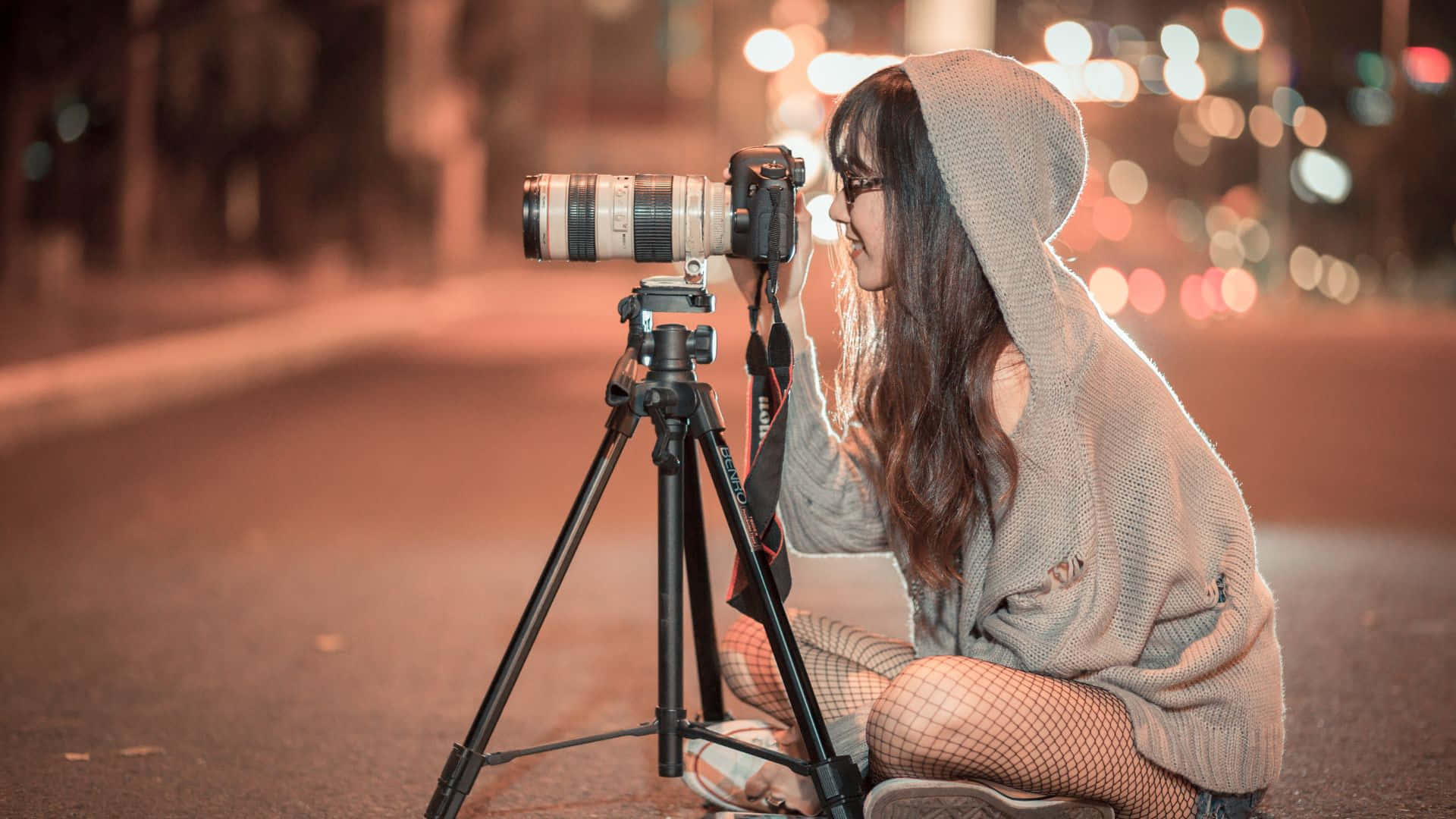 攝影師 桌布 壁紙 — 一位穿著連帽衫的年輕女子在夜晚溫暖的城市燈光下微笑著調整三腳架上的相機。