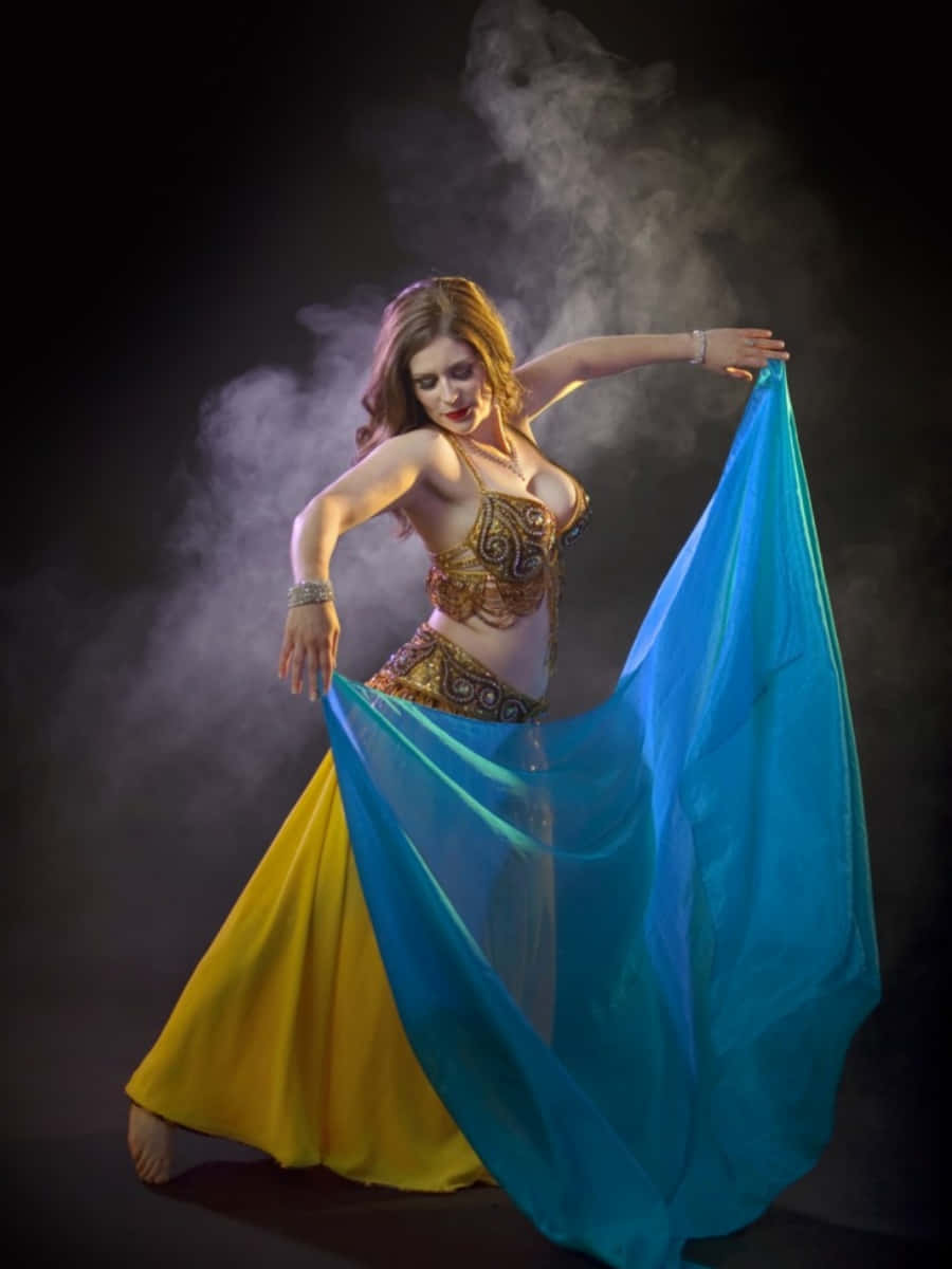 dancer 桌布 壁紙 — 一位穿著金色裝飾服裝的肚皮舞者，在煙霧繚繞的黑暗背景下優雅地手持飄逸的藍色面紗。