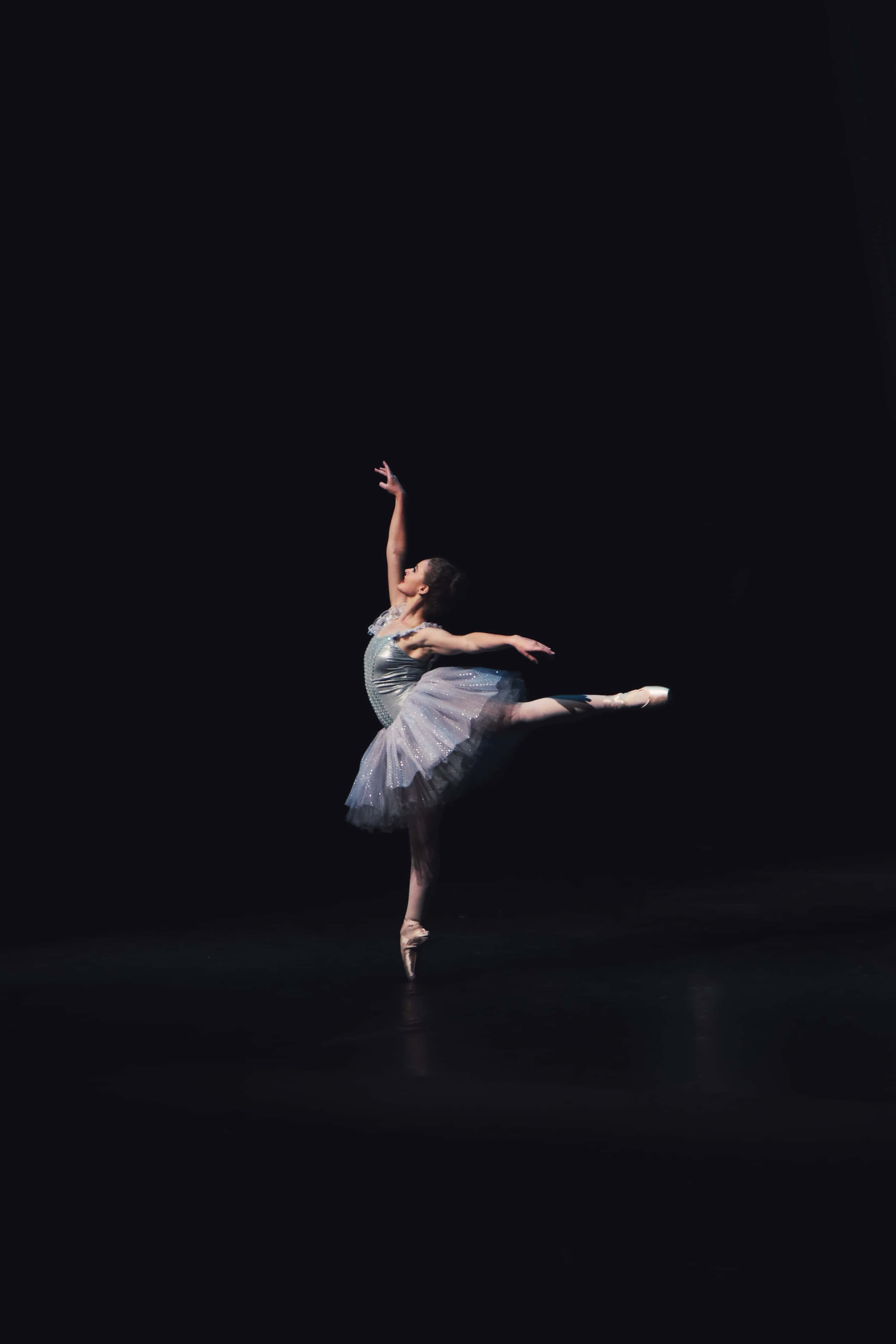 Ballet 桌布 壁紙 — 一位穿著銀色芭蕾舞裙的芭蕾舞者，在強烈聚光燈下，以純黑背景襯托，展現優雅的阿拉貝斯克舞姿。