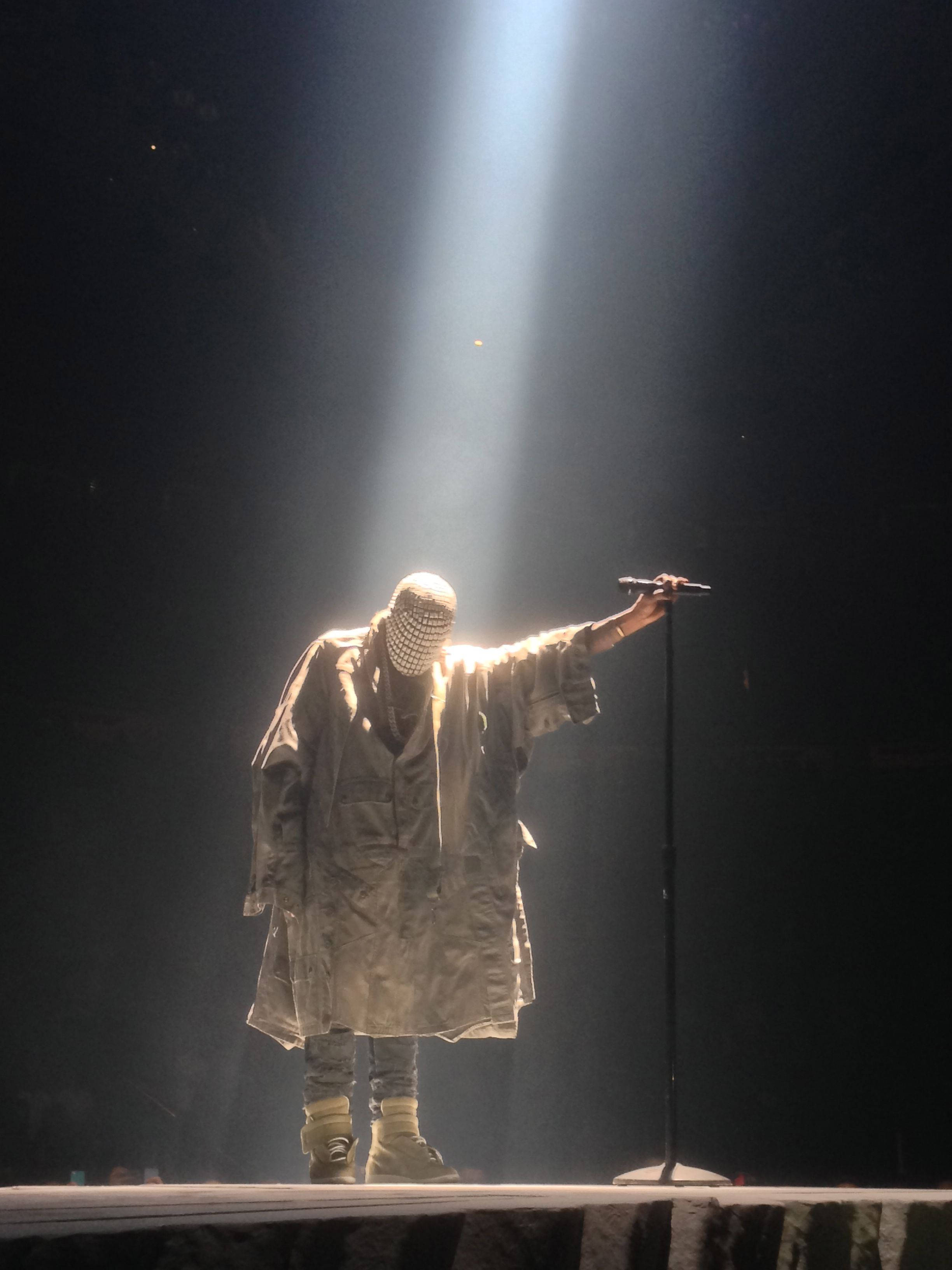 Kanye West Ye 桌布 壁紙 — 一位穿著長外套和圖案面具的表演者站在黑暗的舞台上，沐浴在單一聚光燈下，手持麥克風。