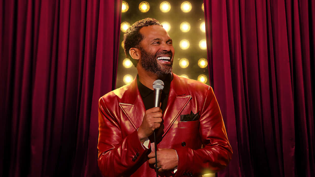 Mike Epps 桌布 壁紙 — 一位穿著閃亮紅色皮夾克的喜劇演員笑著在舞台上拿著麥克風，背景是栗色窗簾和閃爍的聚光燈。
