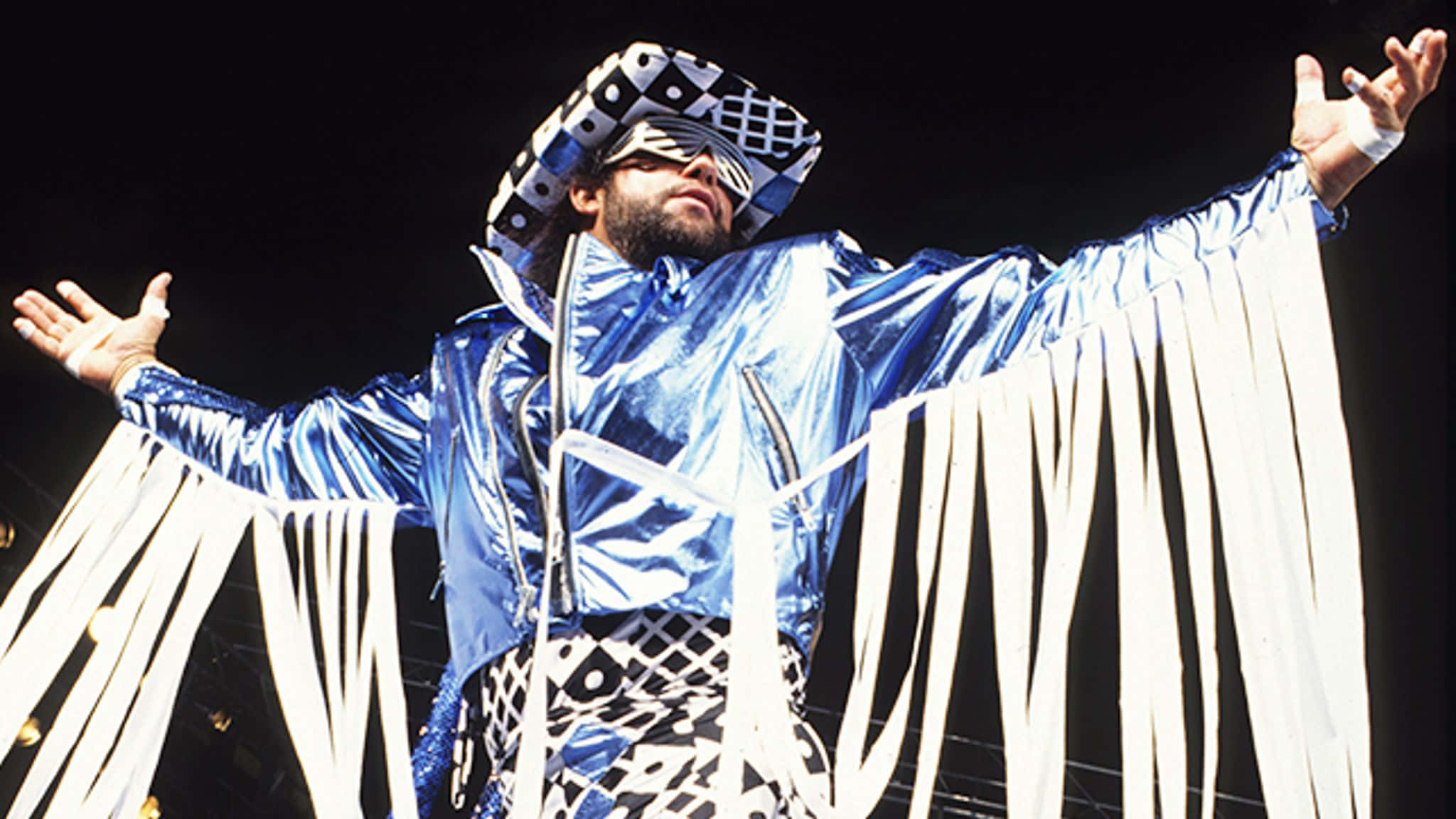 WWE 桌布 壁紙 — 一位穿著閃亮藍色外套和黑白圖案帽子的摔角手，在黑暗的舞台上張開雙臂站立。