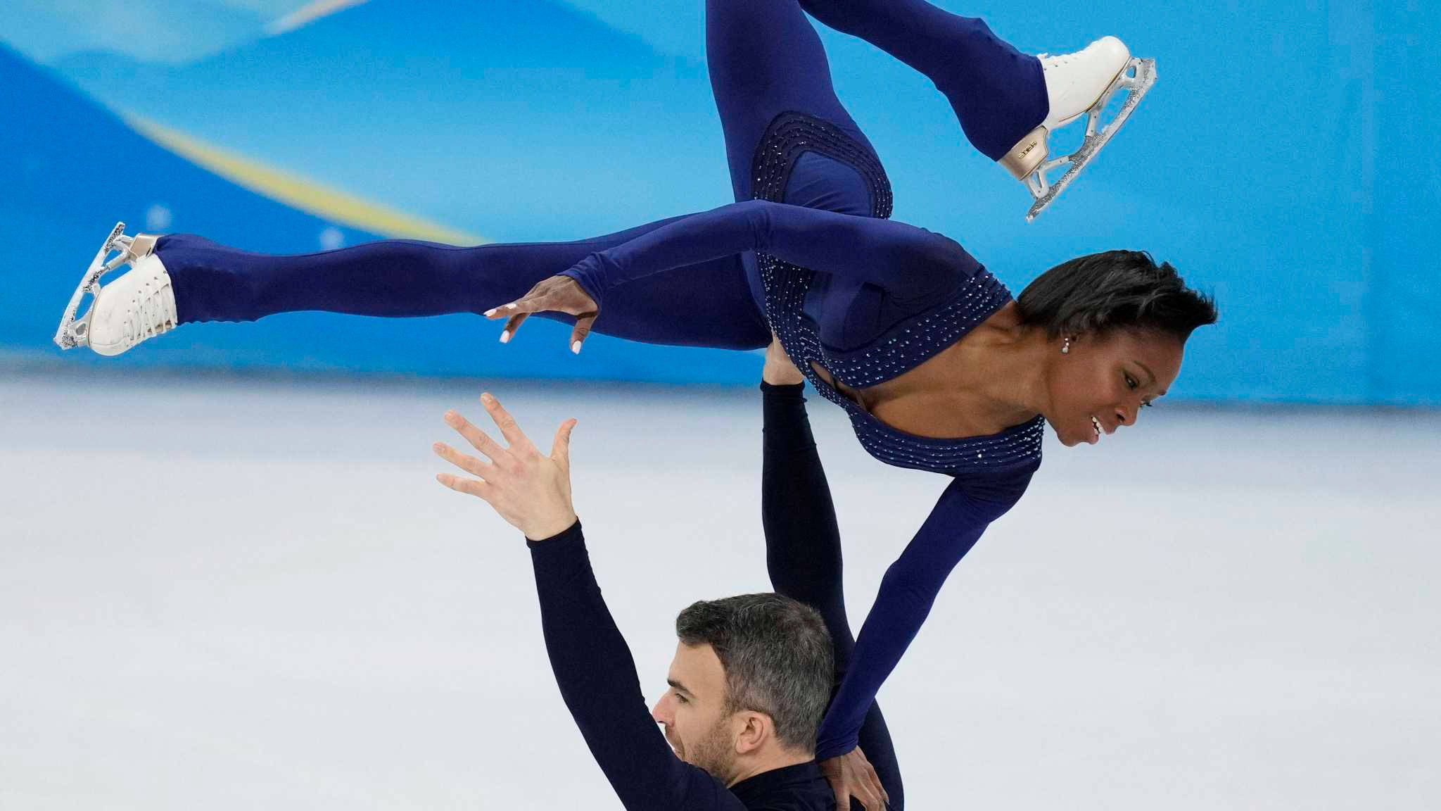 skate 桌布 壁紙 — 一位穿著閃亮藍色服裝的花式滑冰選手在同步表演中被搭檔高高舉起在冰面上。