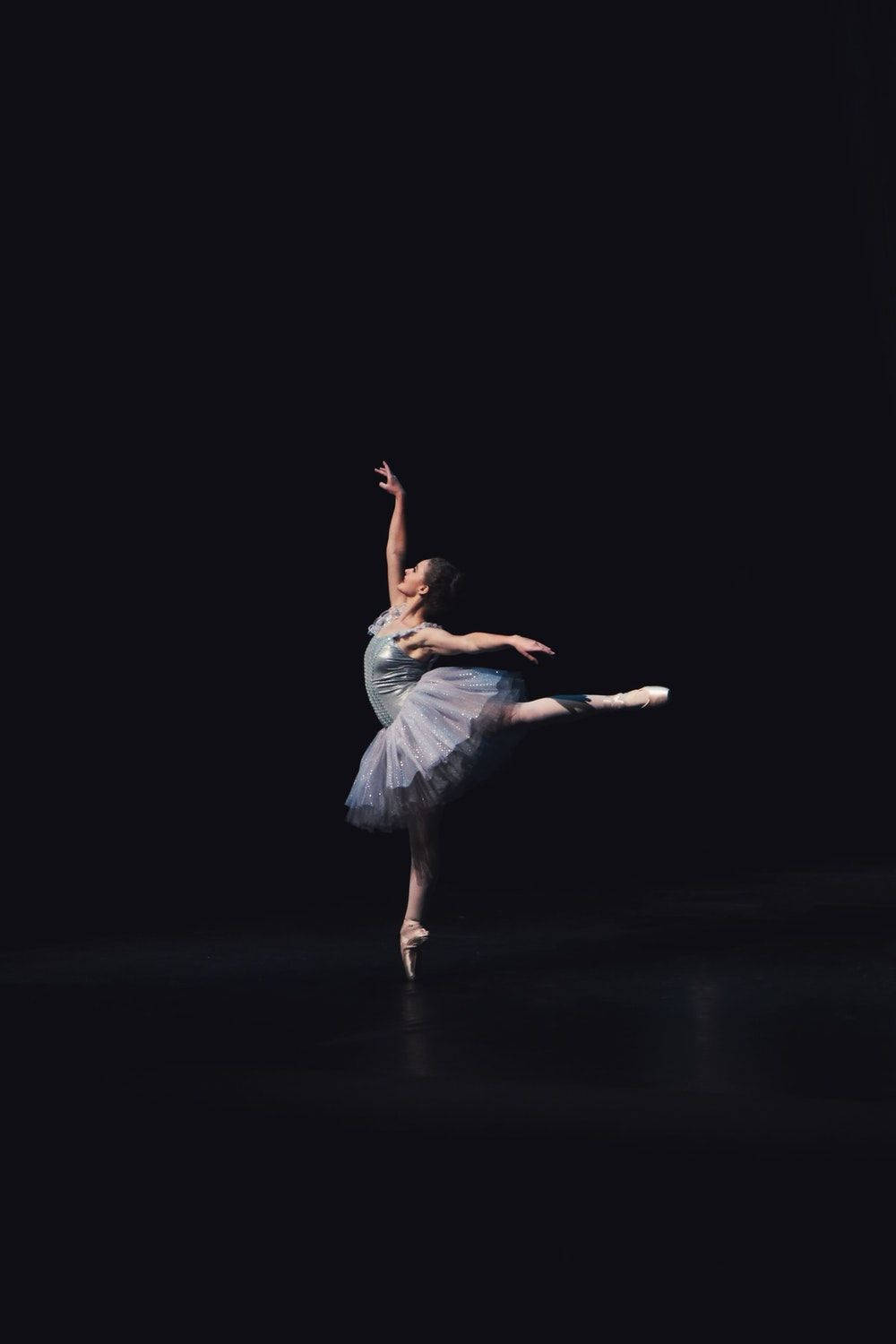 Ballet 桌布 壁紙 — 一位穿著閃亮銀色芭蕾舞裙的芭蕾舞者在強烈的舞台燈光下，在純黑背景前表演優雅的阿拉貝斯克舞姿。