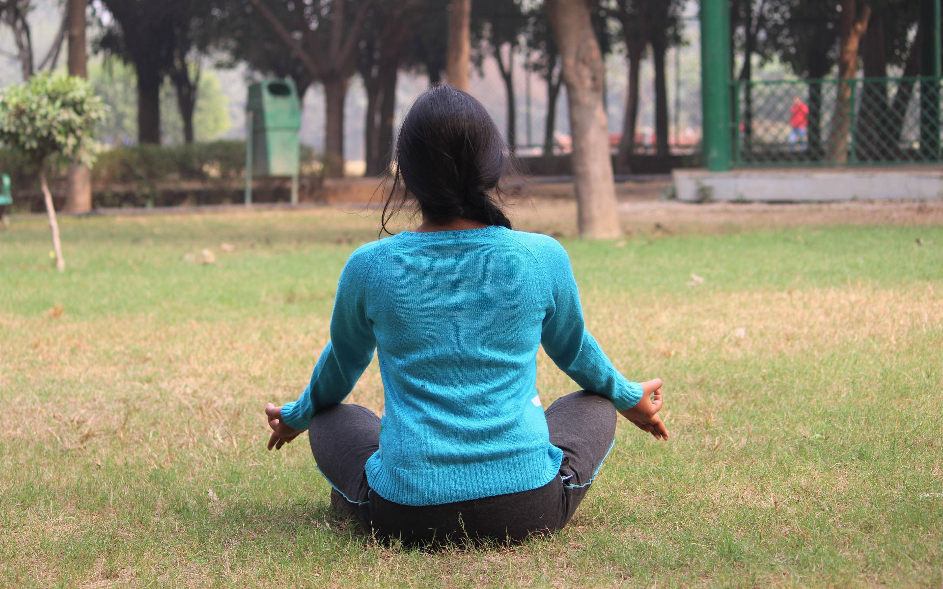 瑜珈 桌布 壁紙 — 一位穿著青綠色毛衣的女性在公園草地上盤腿而坐，背對鏡頭冥想。