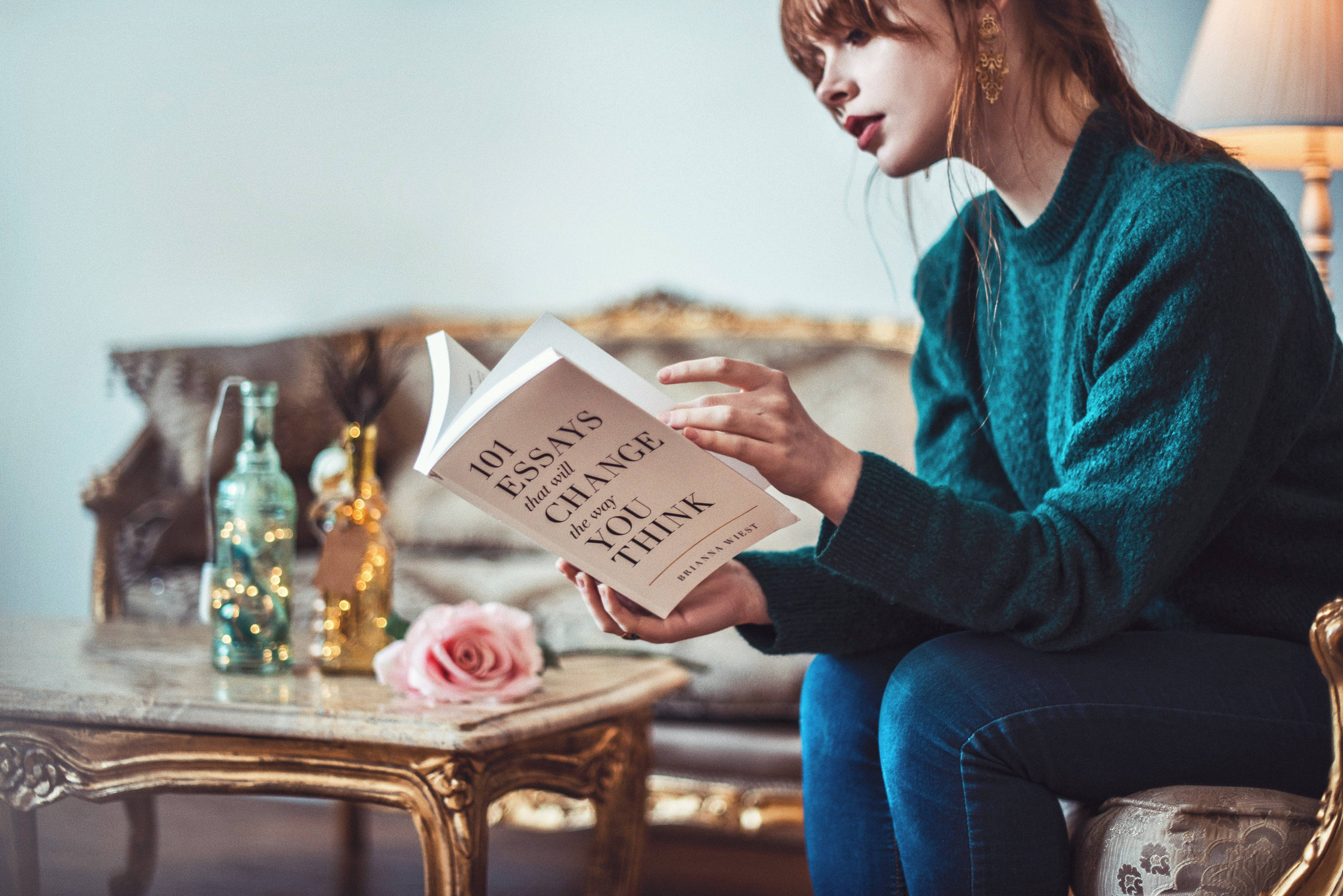 看書 桌布 壁紙 — 一位穿著青綠色毛衣的年輕女性坐在溫暖燈光的復古室內，閱讀一本名為《101 Essays That Will Change the Way You Think》的書。
