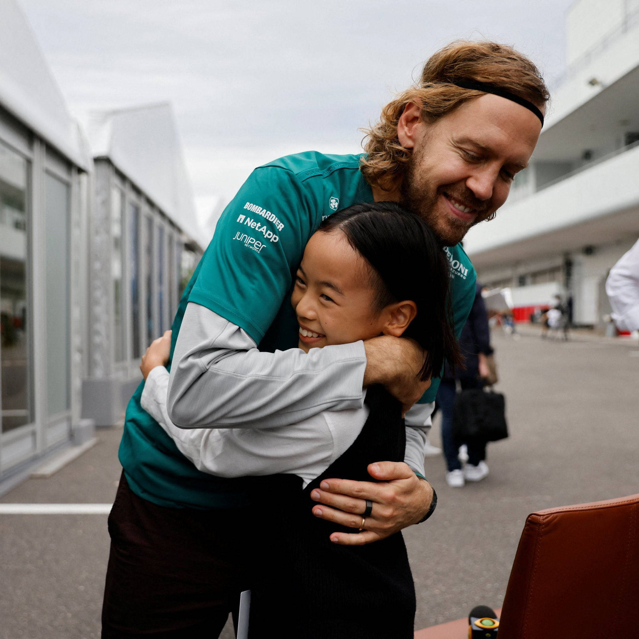sebastian vettel 桌布 壁紙 — 一位穿著青綠色賽車隊襯衫的微笑男子，在賽車場的維修區熱情地擁抱著一個小女孩，充滿喜悅。