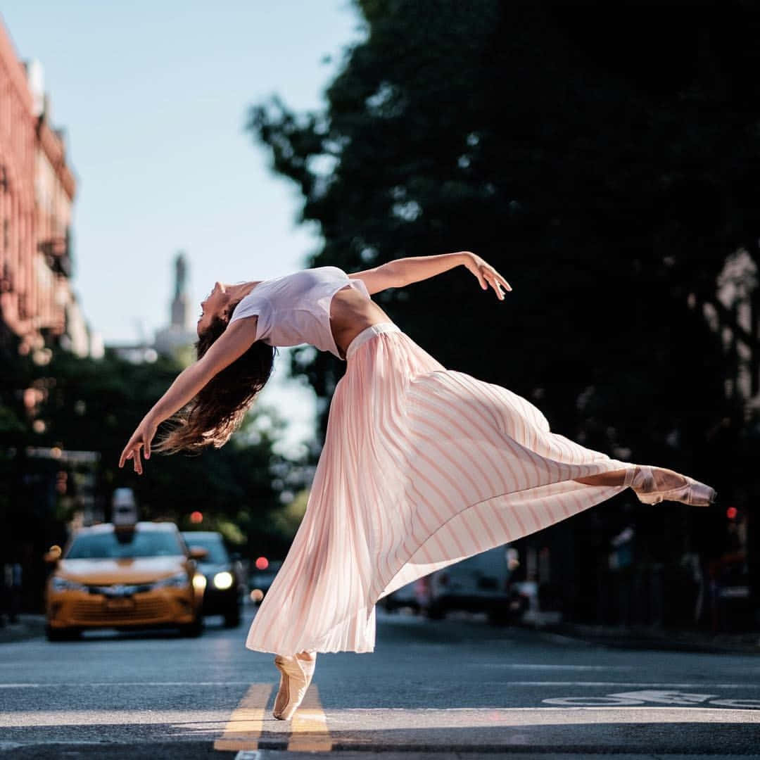 dancer 桌布 壁紙 — 一位穿著飄逸粉紅色裙子的舞者在城市街道上優雅地跳躍，背景有黃色計程車和磚造建築。