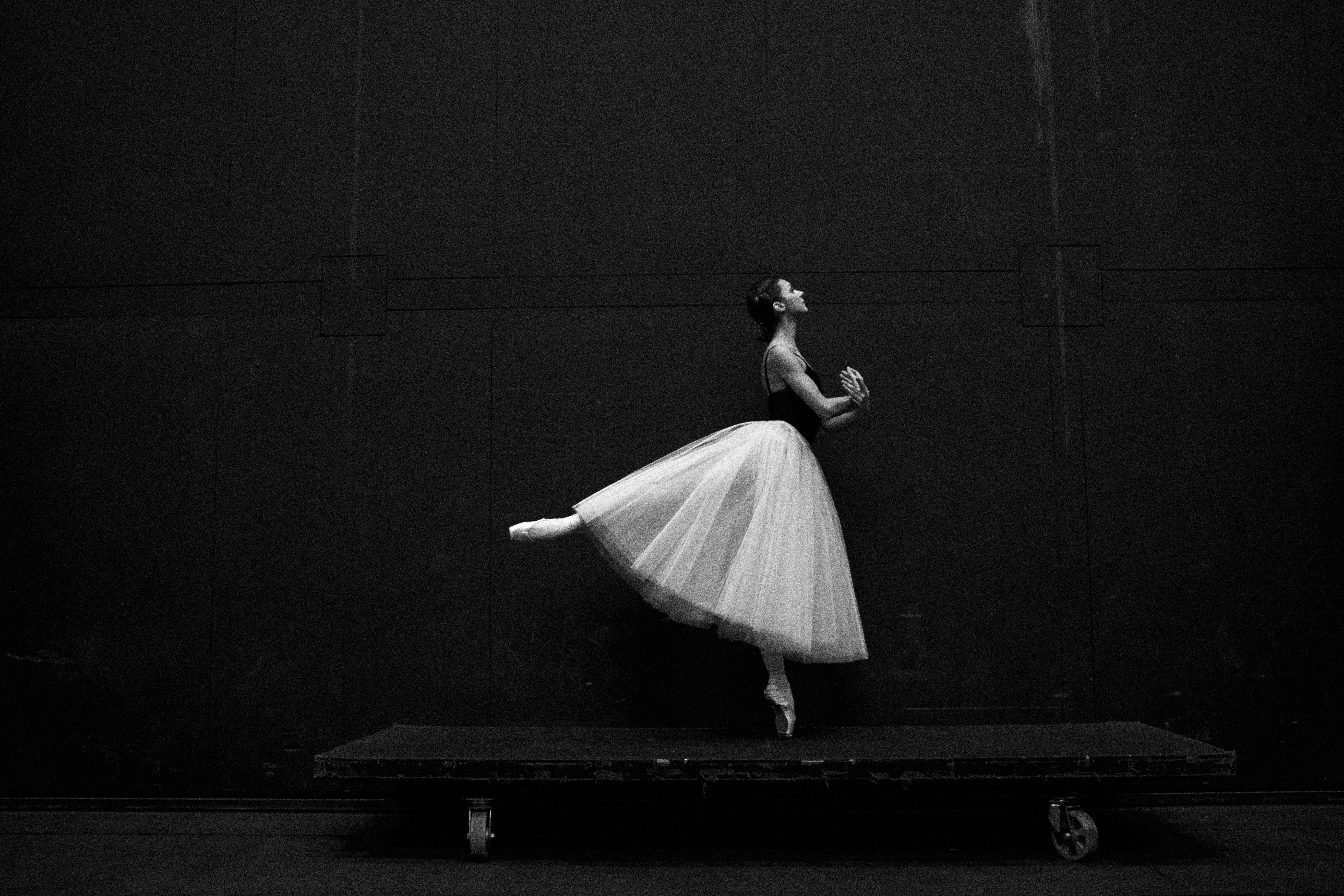 Ballet 桌布 壁紙 — 一位穿著飄逸芭蕾舞裙的芭蕾舞者，在深色牆壁前的輪式舞台上優雅地踮起腳尖保持平衡。