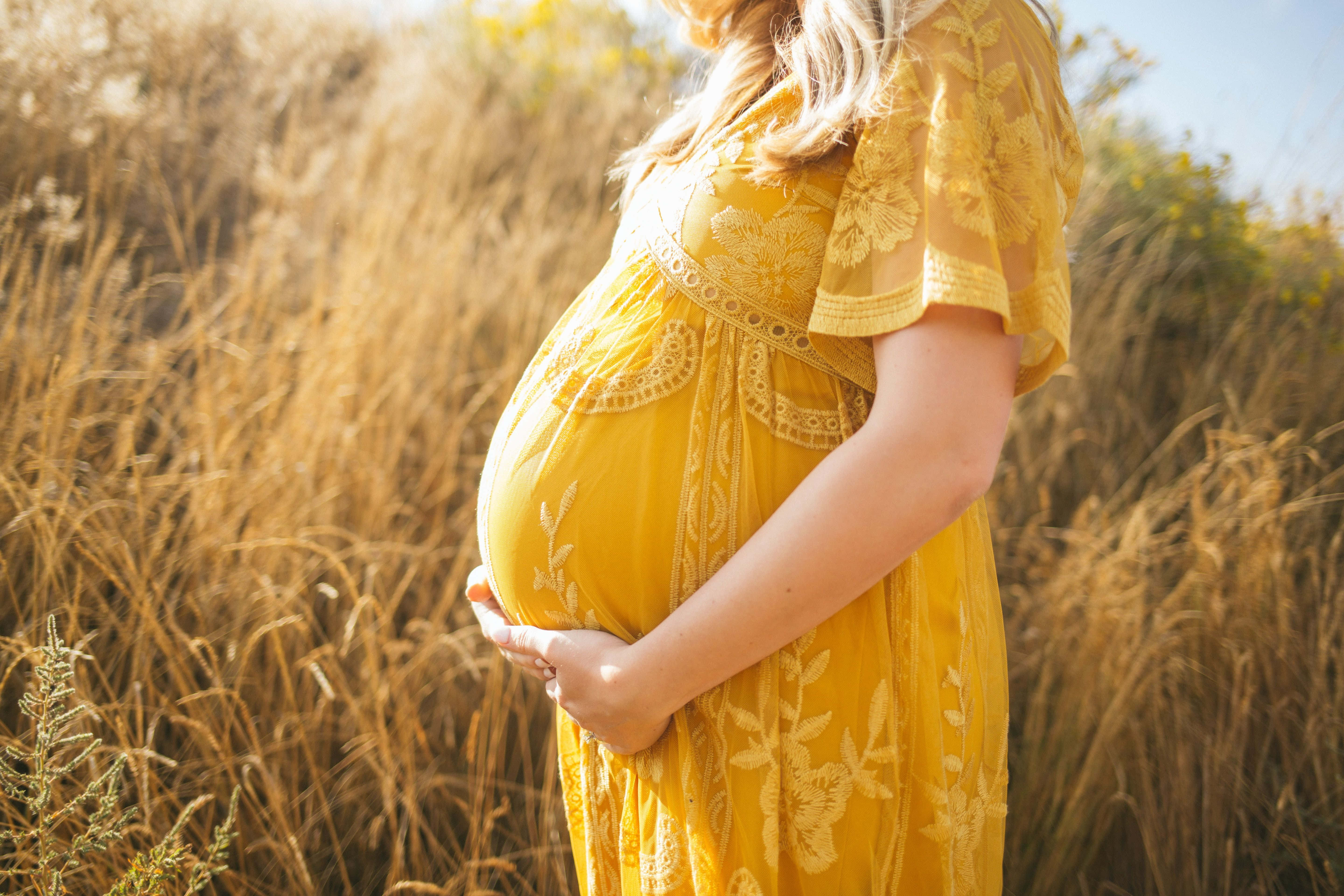 pregnancy 桌布 壁紙 — 一位穿著飄逸黃色連衣裙的孕婦，在金色陽光照耀的草叢中輕撫著她的肚子。