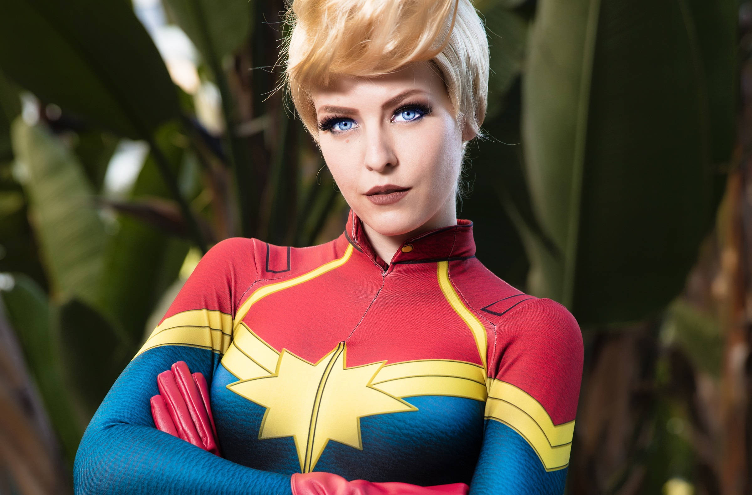 captain marvel 4K 桌布 壁紙 — 一位穿著驚奇隊長服裝的角色扮演者自信地交叉雙臂，站在茂密綠色樹葉的背景前。