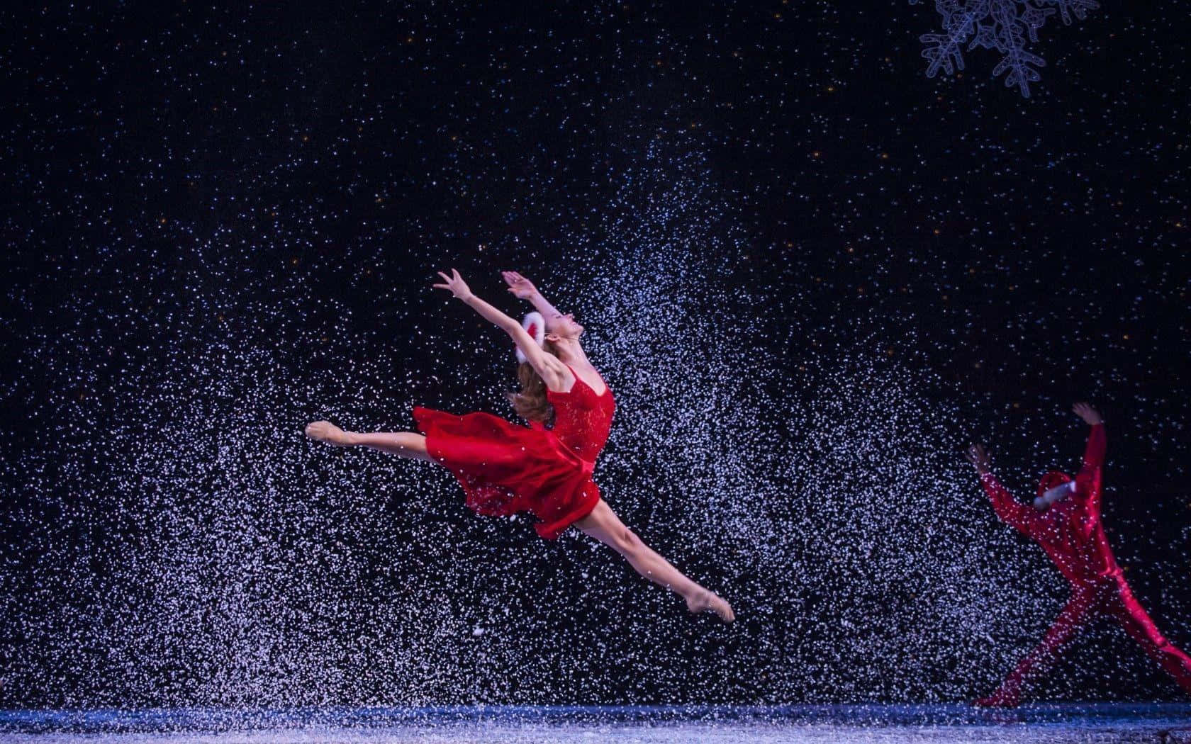Ballet Dancer 桌布 壁紙 — 一位穿著鮮紅色服裝的芭蕾舞者在黑暗舞台背景下，優雅地跳過飄落的雪花。