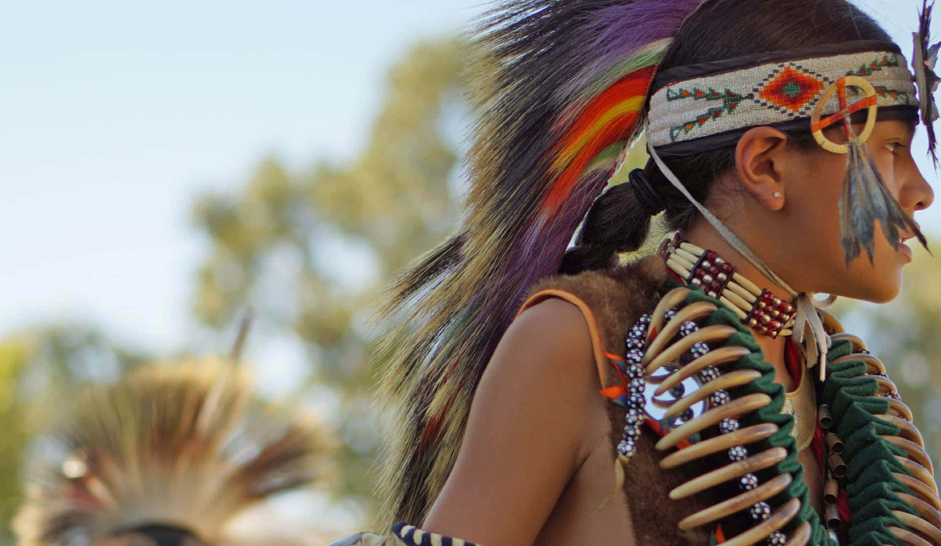Native American 桌布 壁紙 — 一位穿著鮮豔傳統服飾、頭戴羽毛頭飾、頸掛珠鏈的美洲原住民女性，在戶外若有所思地凝視著。
