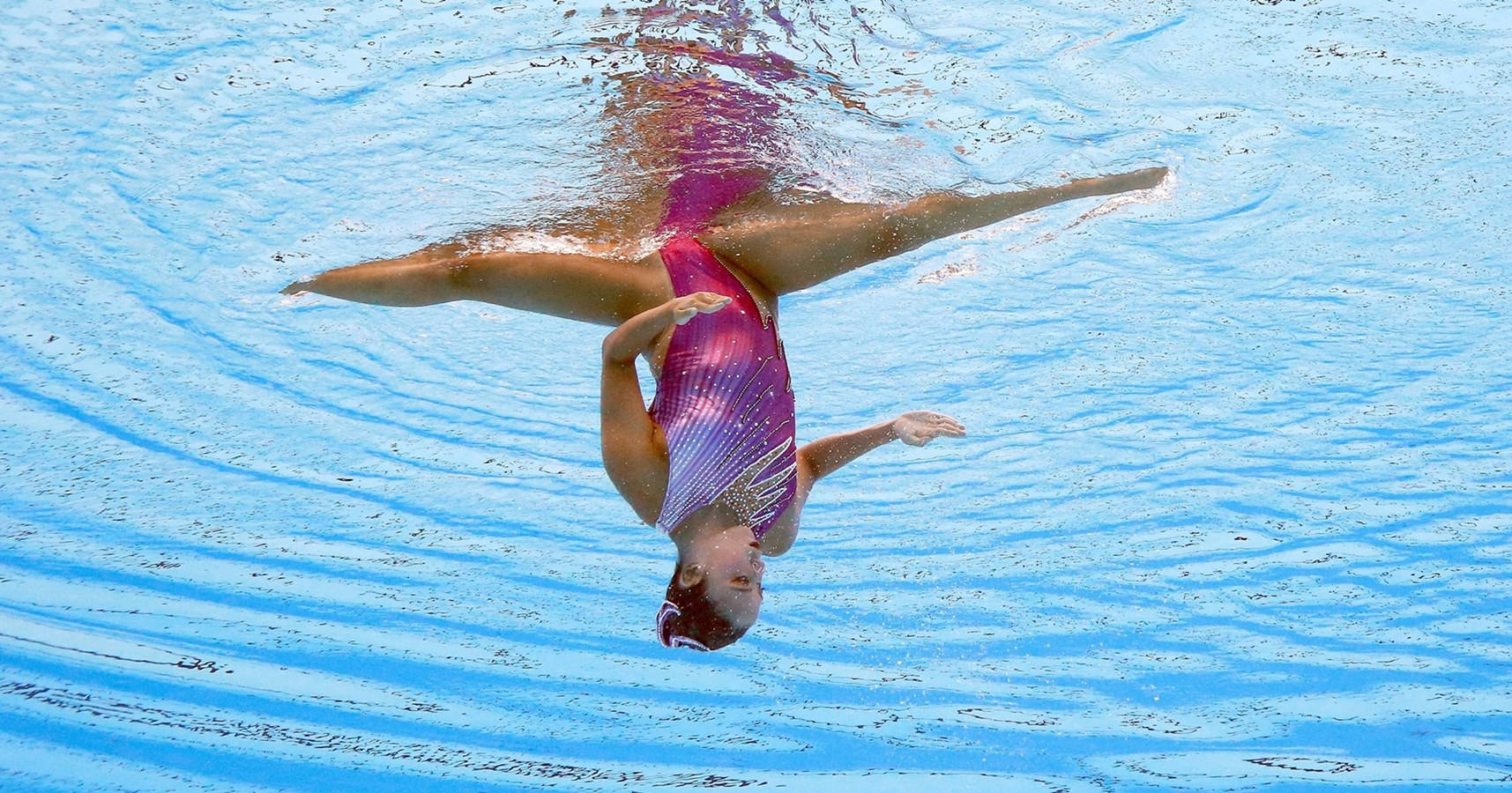 Swimming 桌布 壁紙 — 一位穿著鮮豔紫色泳衣的韻律泳選手在波光粼粼的藍色水面上優雅地做出倒立姿勢。