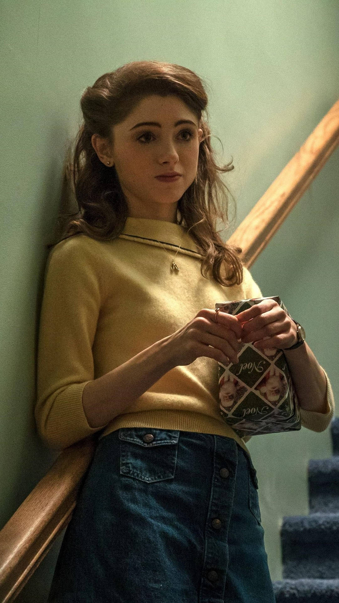 Stranger Things iPhone 桌布 壁紙 — 一位穿著黃色毛衣和牛仔裙的年輕女子靠在樓梯欄杆上，拿著一個圖案包。