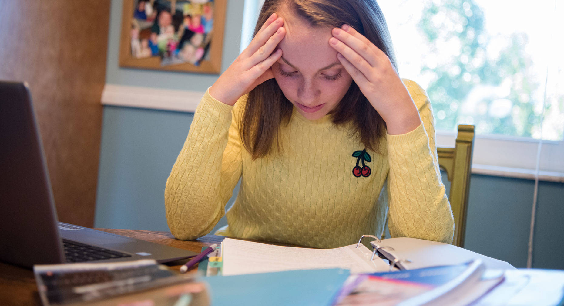 Anxiety 桌布 壁紙 — 一位穿著黃色毛衣的年輕女性，在雜亂的書桌前用筆記型電腦和書籍學習時，沮喪地抱著頭。