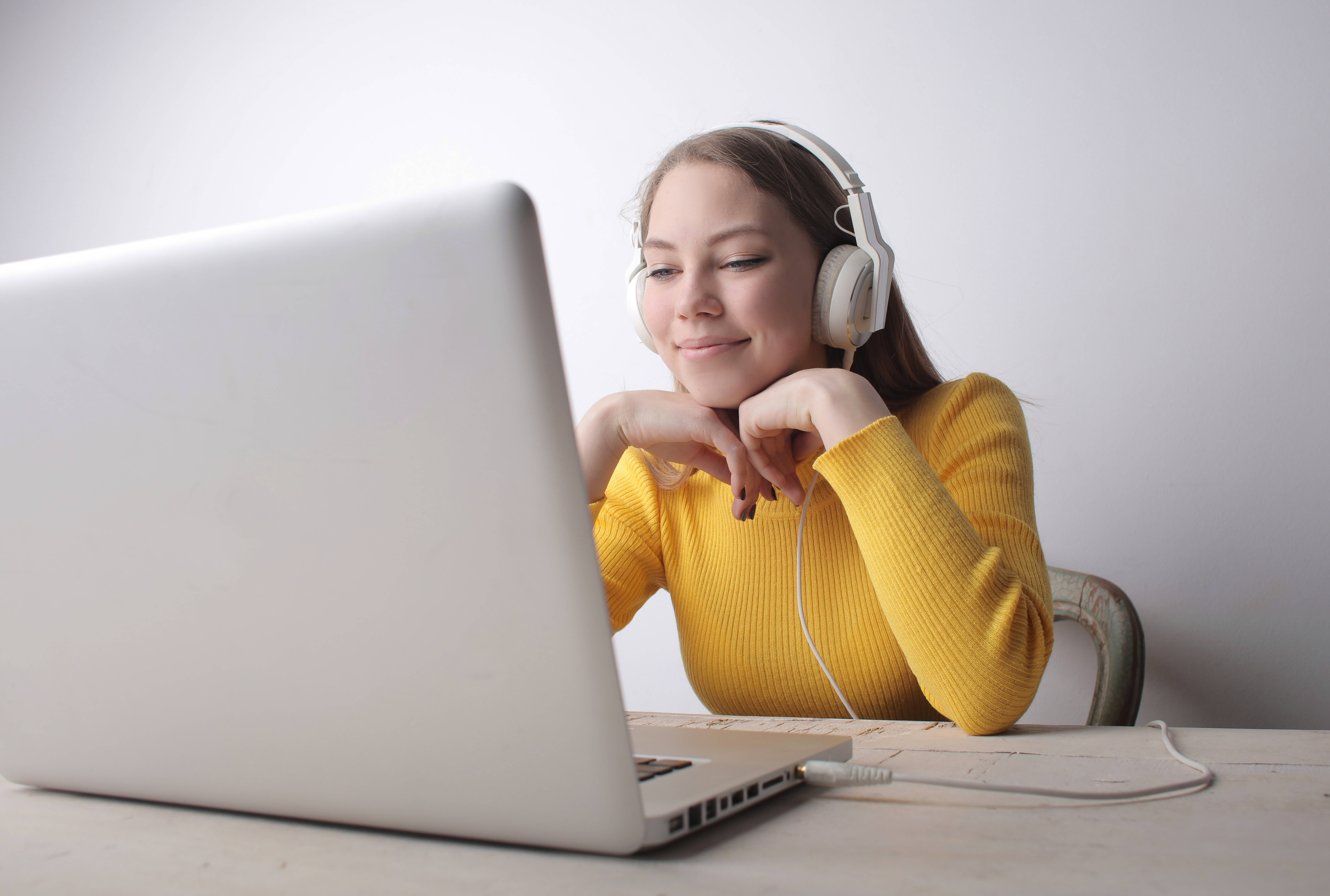 Cute Laptop 桌布 壁紙 — 一位穿著黃色毛衣的年輕女性微笑著，戴著白色耳機，在木桌上使用筆記型電腦。