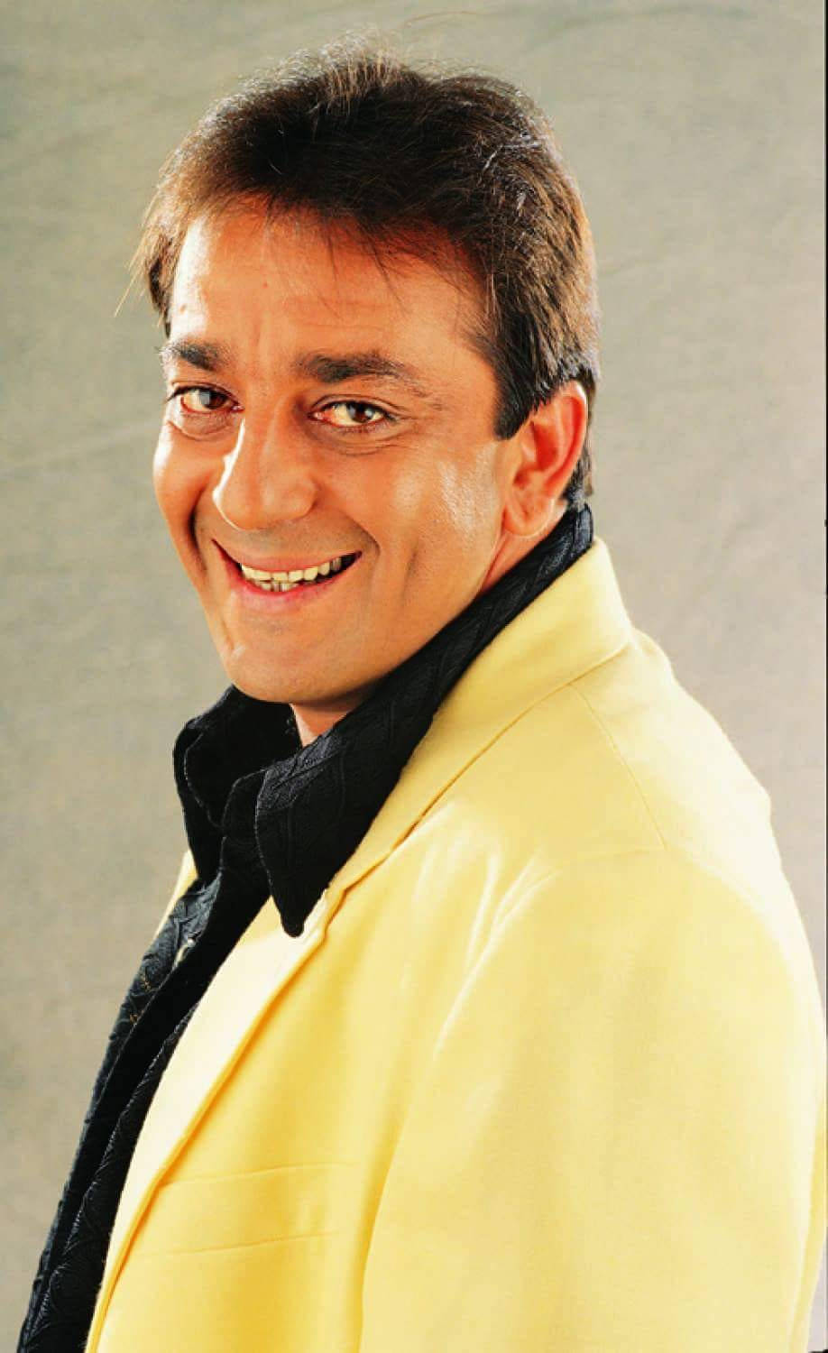 Sanjay 桌布 壁紙 — 一位穿著黃色西裝外套的開朗男士，在灰色中性攝影棚背景前溫暖地微笑。