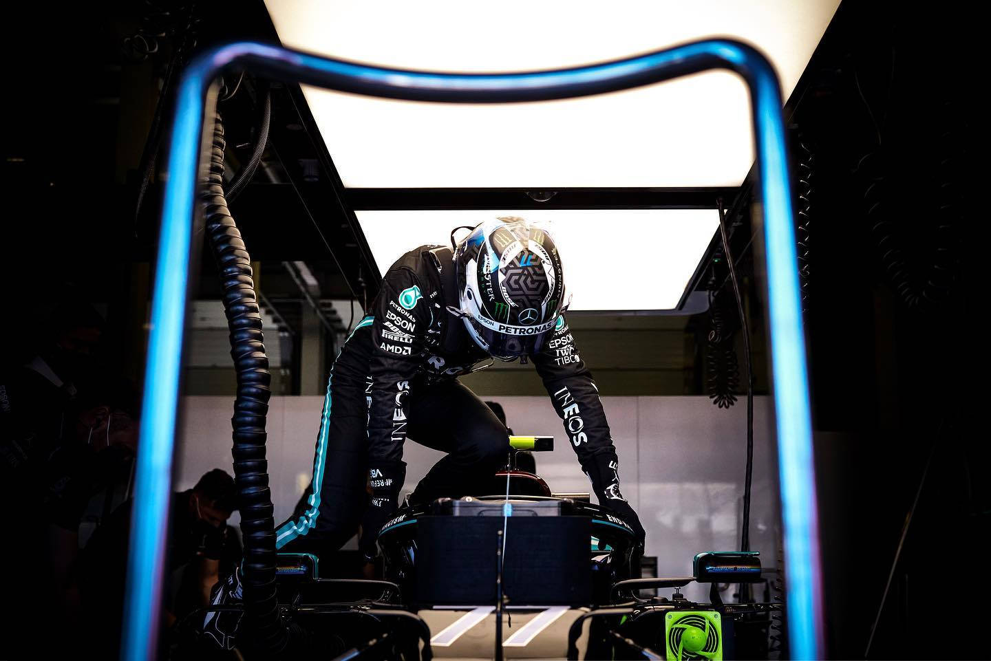 Valtteri Bottas 桌布 壁紙 — 一位穿著黑色和青綠色賽車服的F1車手在昏暗的車庫中，在明亮的頭頂燈光下俯身在他的賽車上。