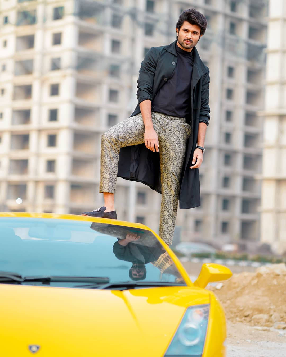 vijay devarakonda 4K 桌布 壁紙 — 一位穿著黑色外套和圖案褲子的時尚男士，自信地站在一輛亮黃色跑車上，背景是未完工的高樓大廈。