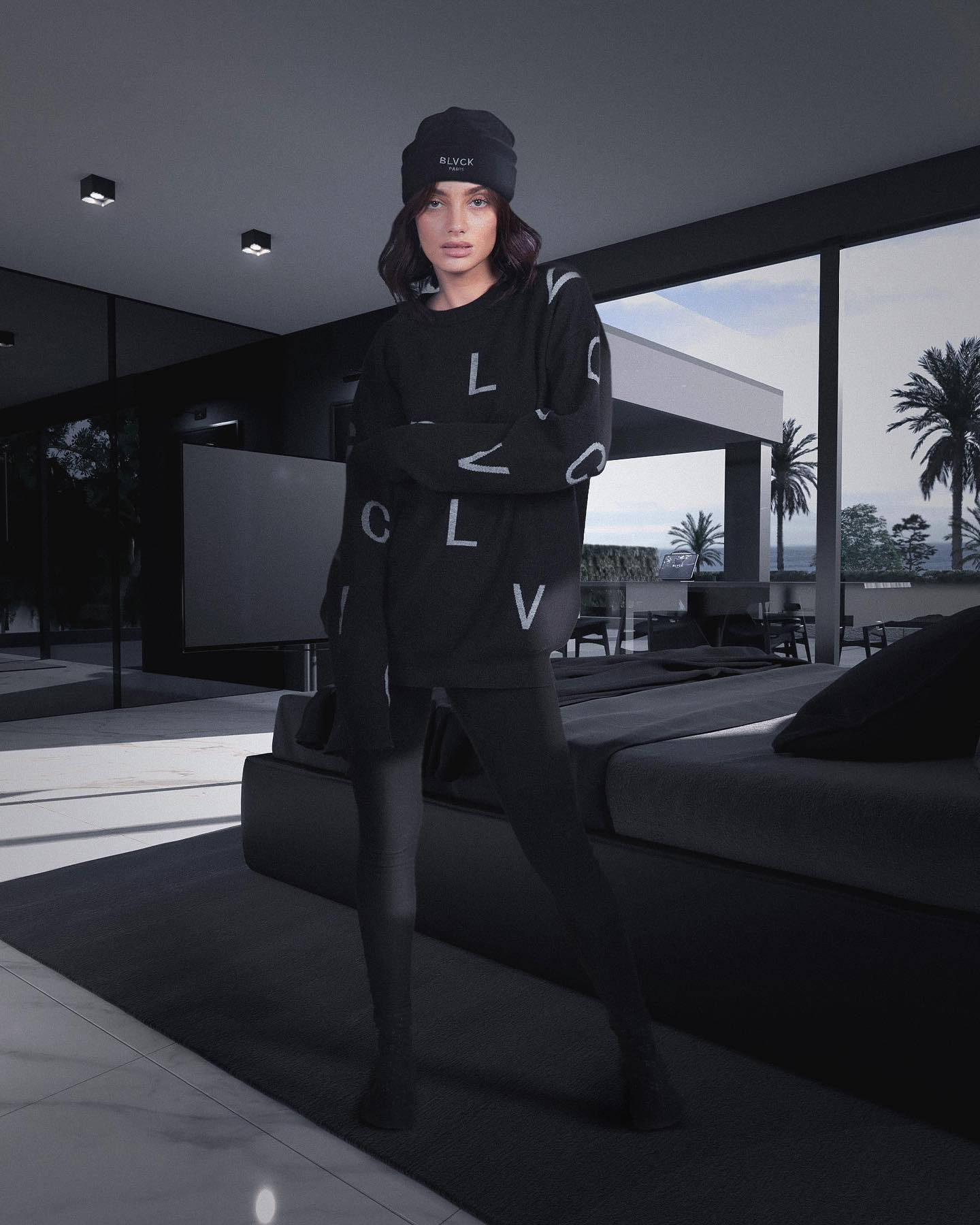 Blvck Paris 桌布 壁紙 — 一位穿著黑色字母組合休閒服的女人自信地站在一個極簡現代住宅中，落地窗俯瞰棕櫚樹。