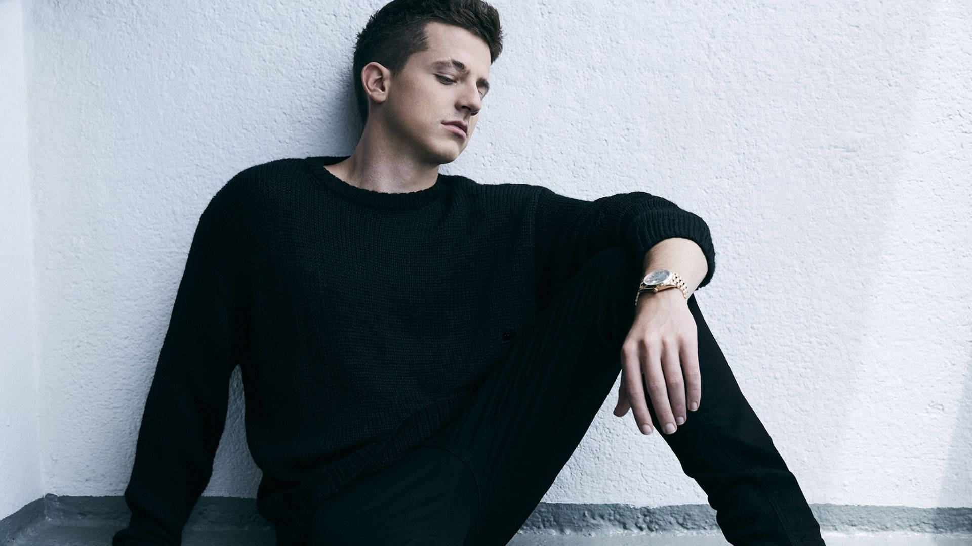 Charlie Puth 桌布 壁紙 — 一位穿著黑色毛衣的年輕男子靠著白色紋理牆，手腕上戴著金色手錶。