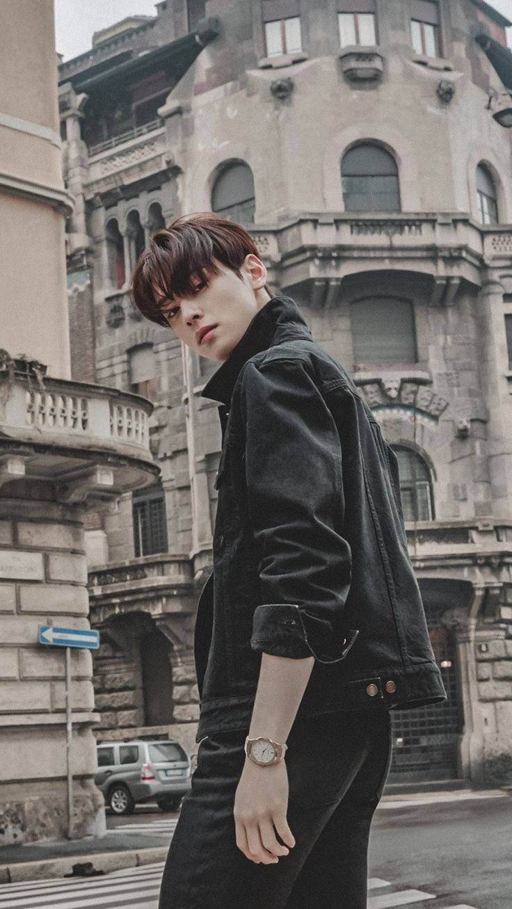 Eunwoo 桌布 壁紙 — 一位穿著黑色牛仔外套的年輕男子自信地站在一條擁有歷史建築的城市街道上。