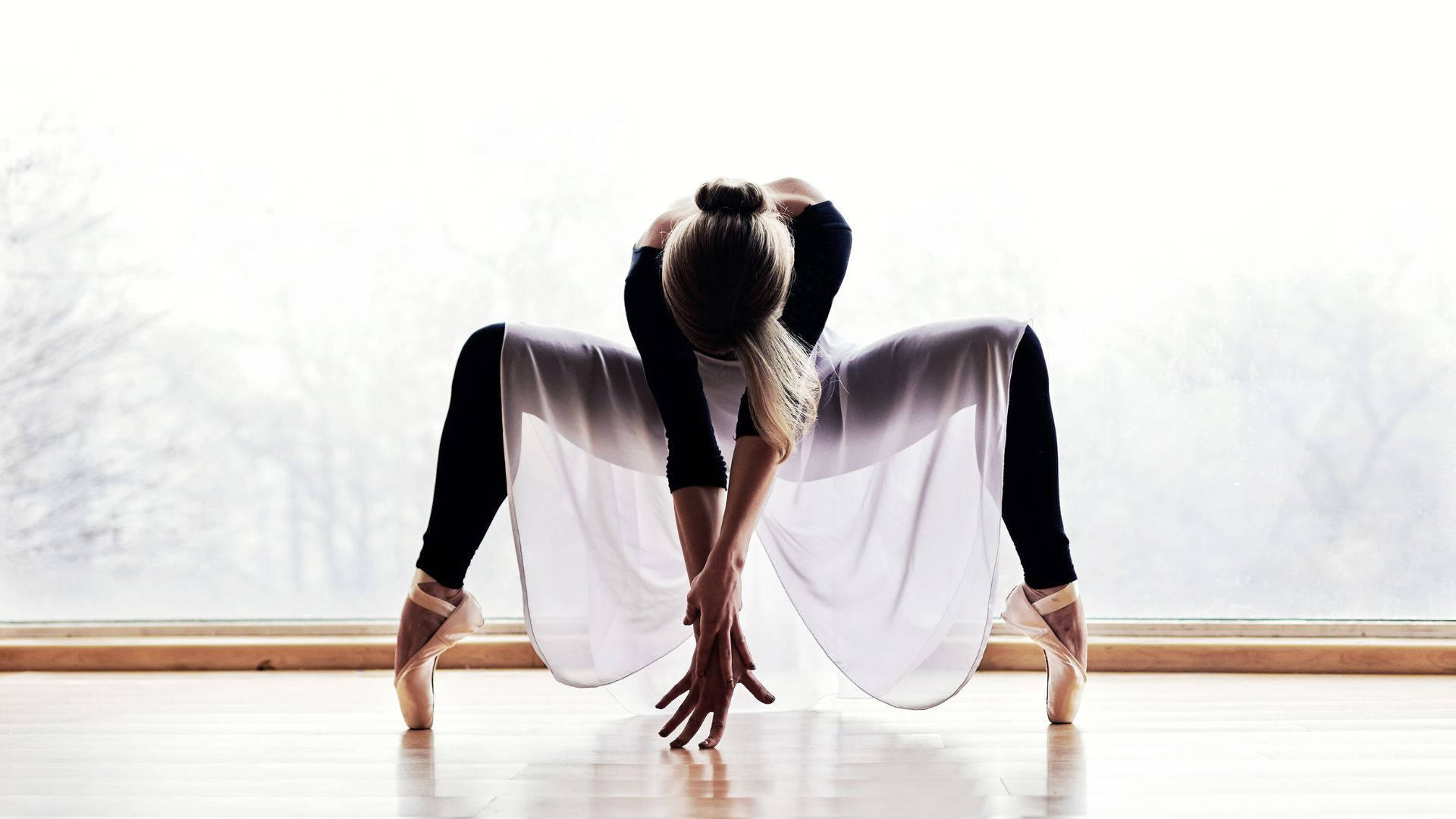 Ballet 桌布 壁紙 — 一位穿著黑色緊身衣和飄逸白色裙子的芭蕾舞者在陽光明媚的練習室中優雅地伸展，窗外可見光禿禿的樹木。