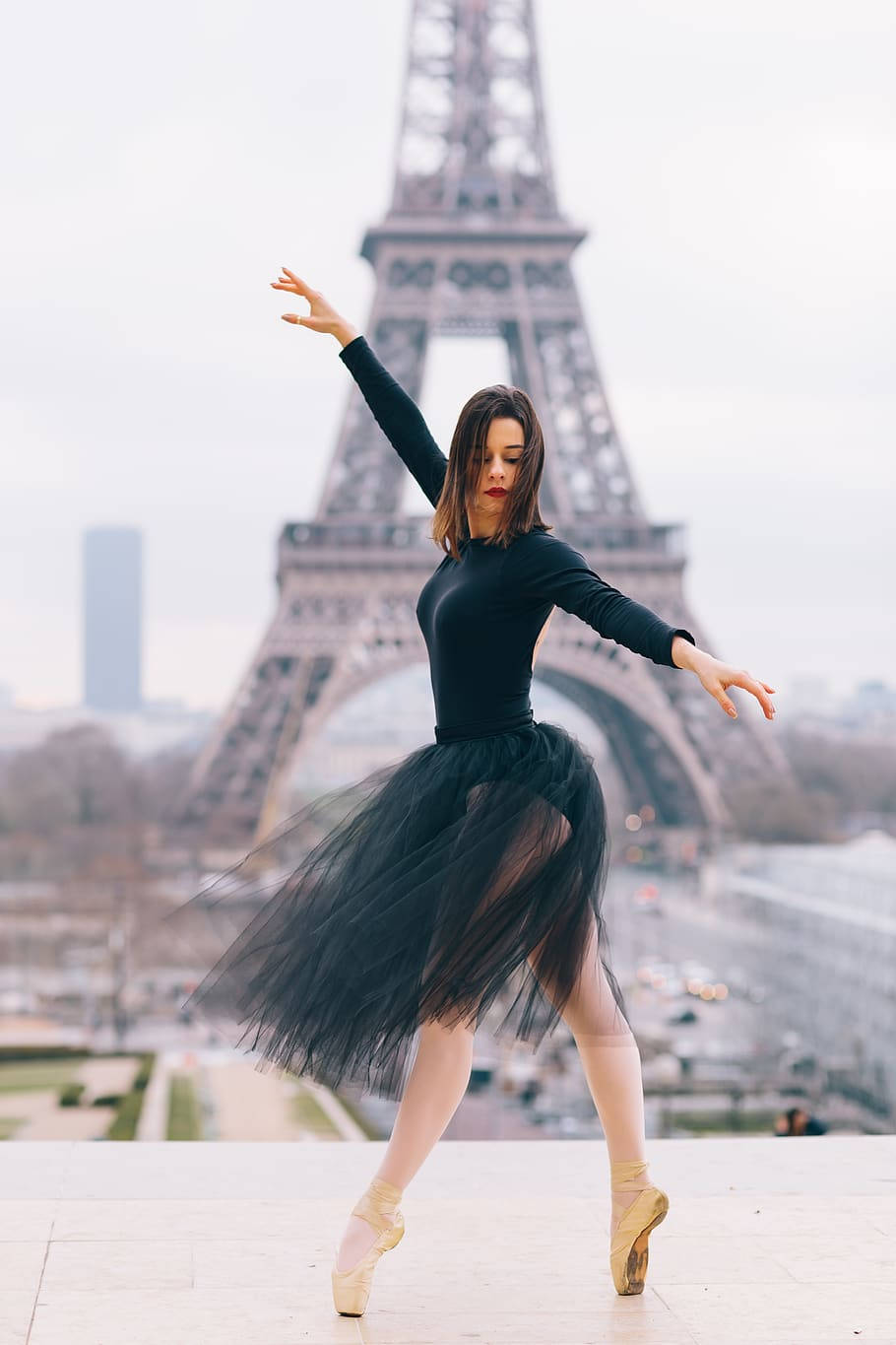 Eiffel Tower 桌布 壁紙 — 一位穿著黑色芭蕾舞裙的芭蕾舞者，在柔和日光下，以足尖優雅地擺出姿勢，背後是巍峨的艾菲爾鐵塔。