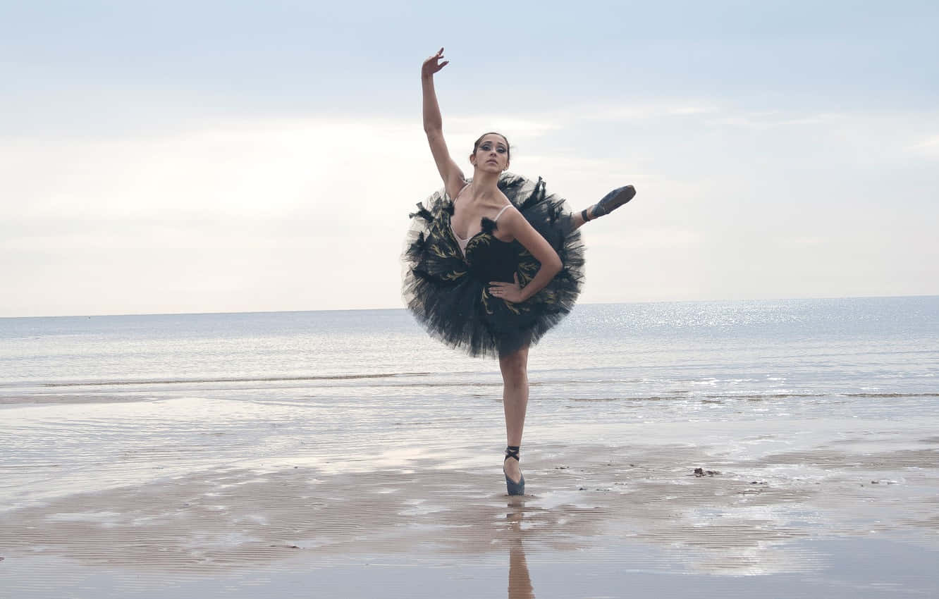 Ballet 桌布 壁紙 — 一位穿著黑色芭蕾舞裙的芭蕾舞者在濕沙灘上擺出優雅的姿勢，身後是海浪和蒼白的天空。