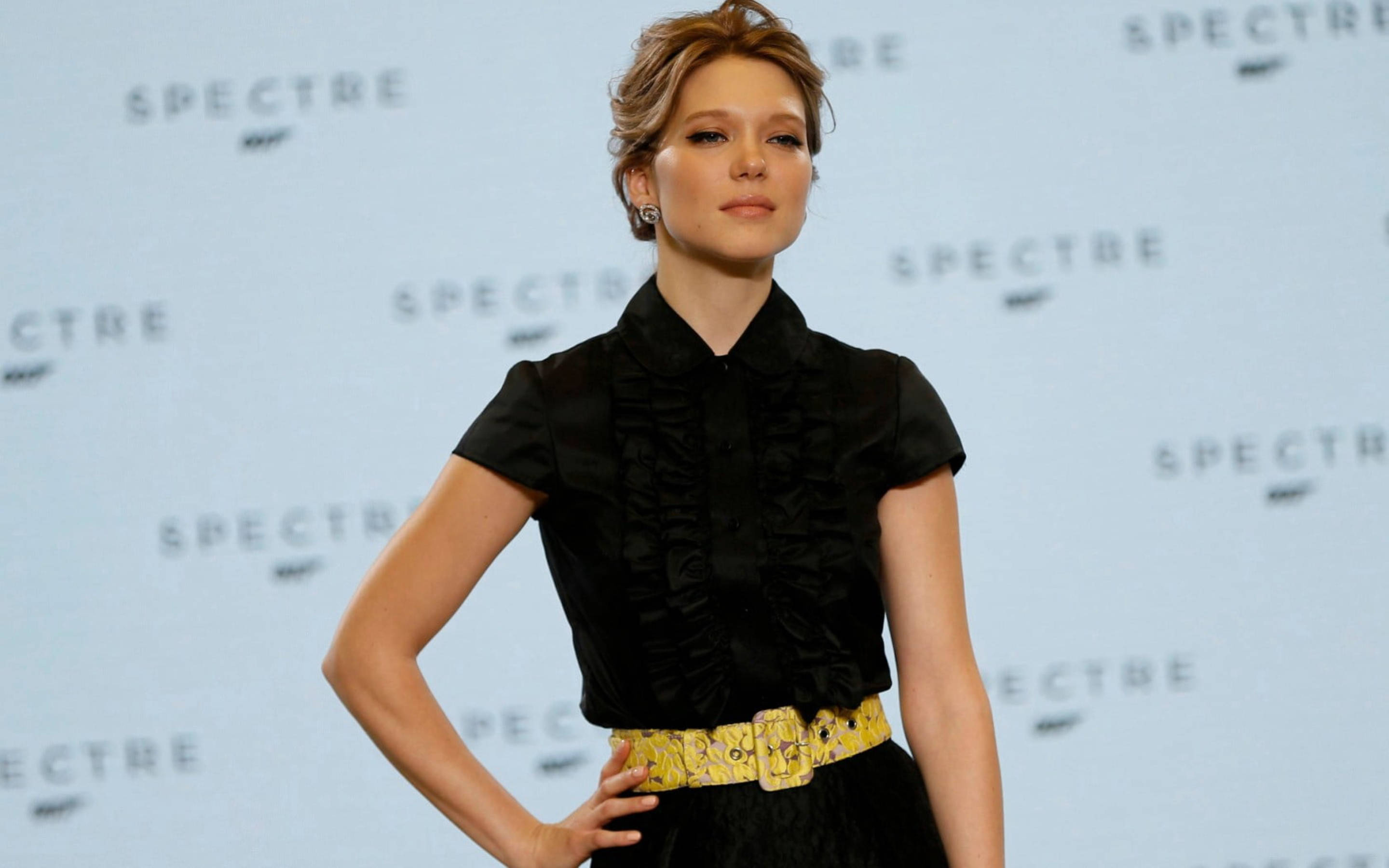 Lea Seydoux 桌布 壁紙 — 一位穿著黑色荷葉邊襯衫和黃色圖案腰帶的優雅女性，自信地站在重複的Spectre背景前。