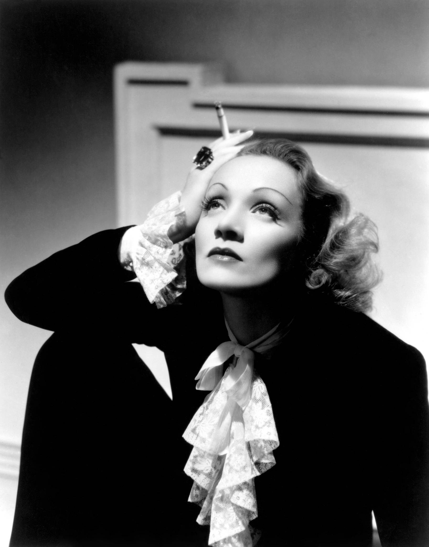 Marlene Dietrich 桌布 壁紙 — 一位穿著黑色西裝、帶有蕾絲袖口和蝴蝶結的迷人女士，在戲劇性的工作室肖像中將香煙靠近太陽穴。