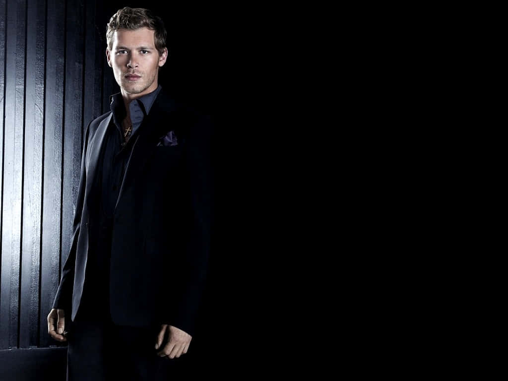 Vampire Diaries 桌布 壁紙 — 一位穿著黑色西裝、打扮俐落的男士，在戲劇性的工作室燈光下，站在深色垂直鑲板牆前。
