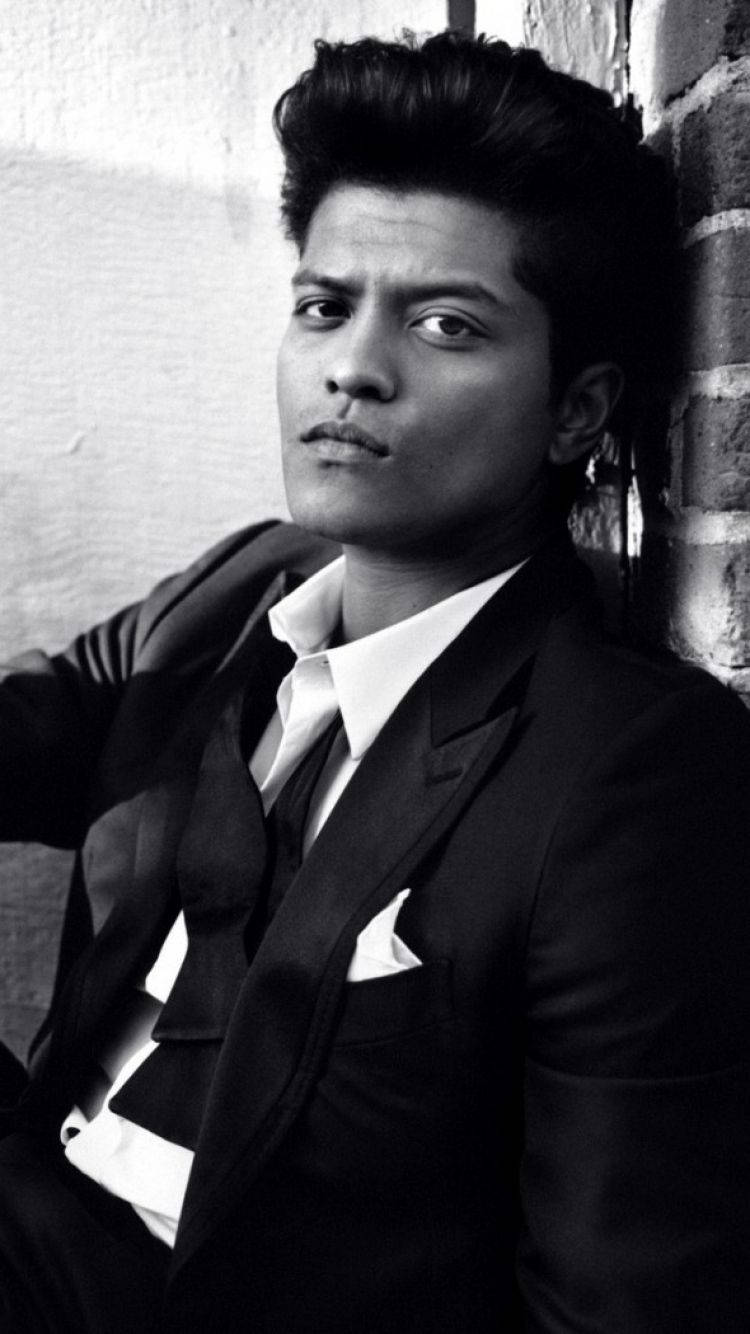 Bruno Mars 桌布 壁紙 — 一位穿著黑色西裝、打扮俐落的男士，在高對比度的黑白肖像中，靠著磚牆，眼神銳利。