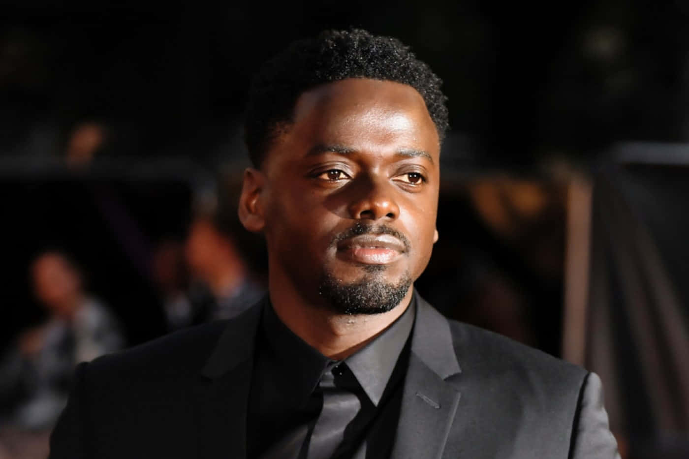Daniel Kaluuya 桌布 壁紙 — 一位穿著黑色西裝、打扮俐落的男士，在昏暗模糊的背景前凝視著，柔和的光線凸顯了他的面部特徵。