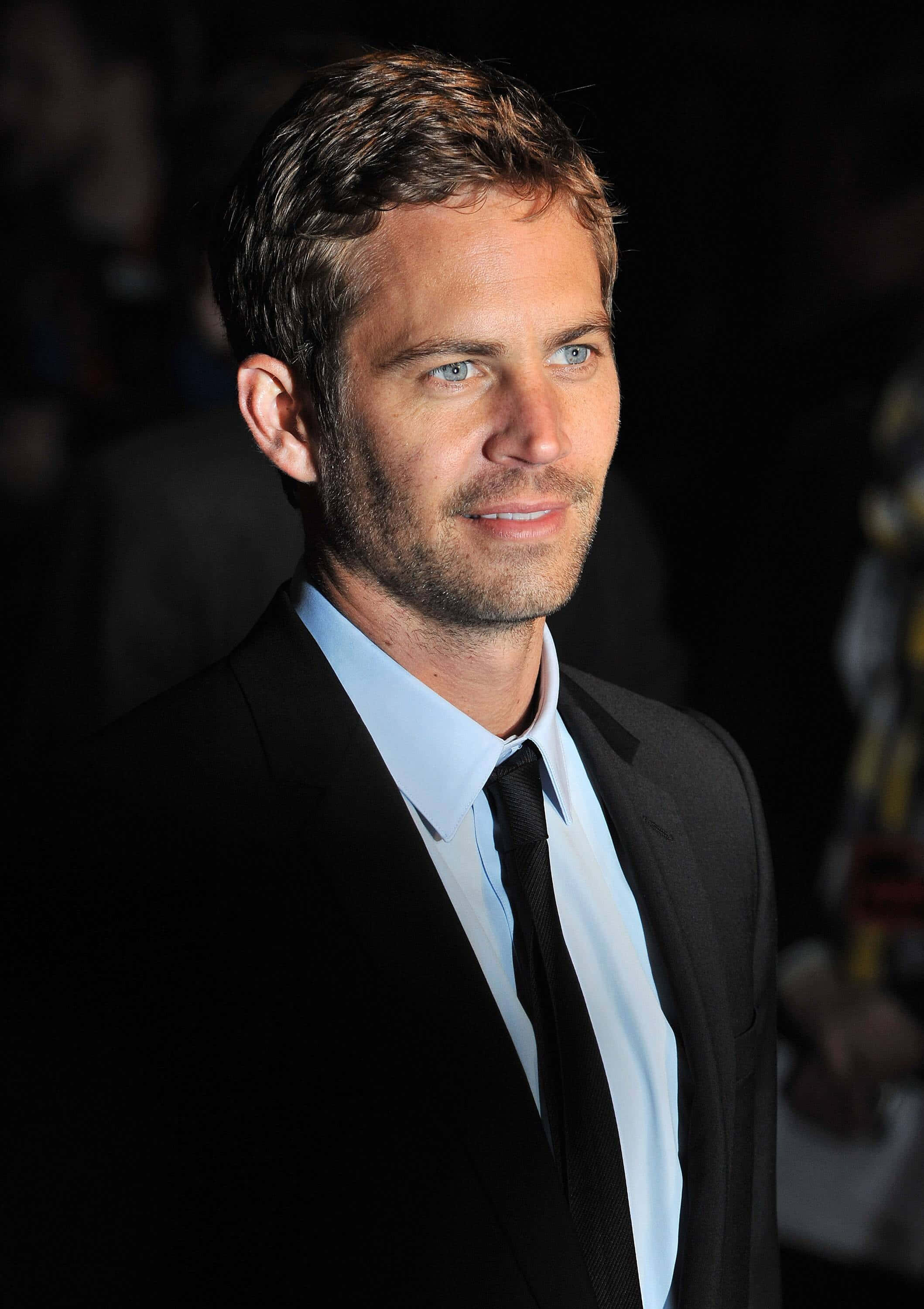 Paul Walker 桌布 壁紙 — 一位穿著黑色西裝和淺藍色襯衫、衣著講究的男子，在黑暗模糊的背景前露出微妙的微笑。