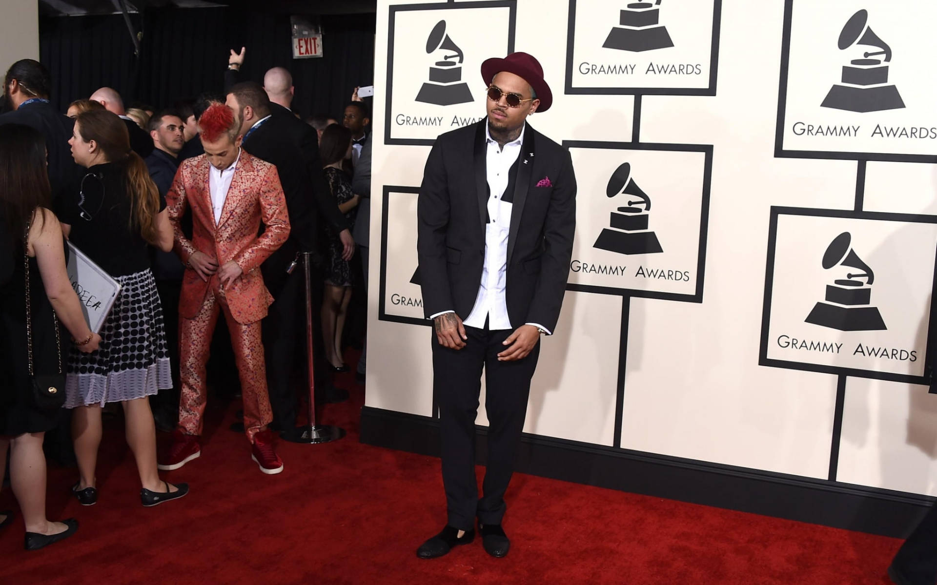 Chris Brown 桌布 壁紙 — 一位穿著黑色西裝和紅色帽子的時尚男士在葛萊美獎紅地毯上自信地擺姿勢，附近有一位穿著閃閃發光金色西裝的人。