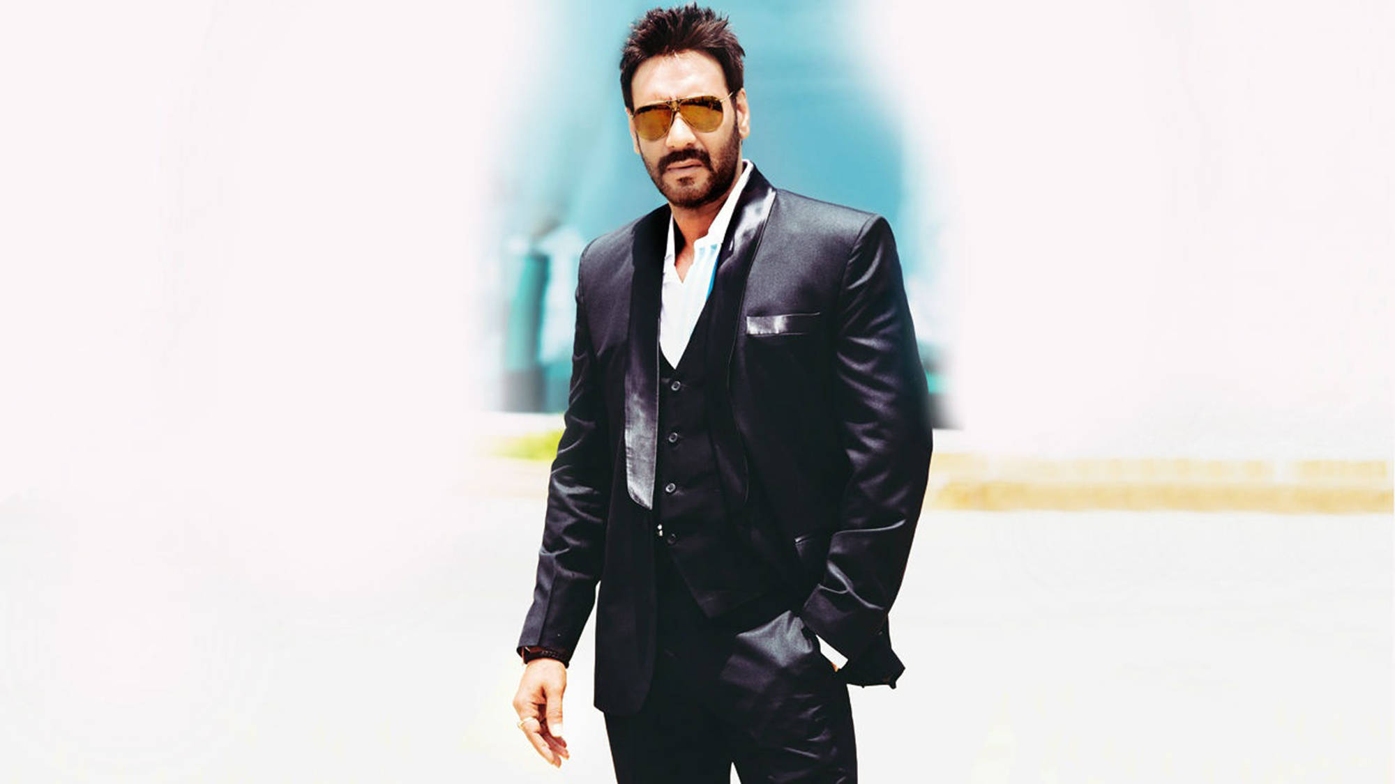 ajay devgn 桌布 壁紙 — 一位穿著黑色西裝和金色太陽眼鏡的時尚男士，自信地站在明亮模糊的背景前。
