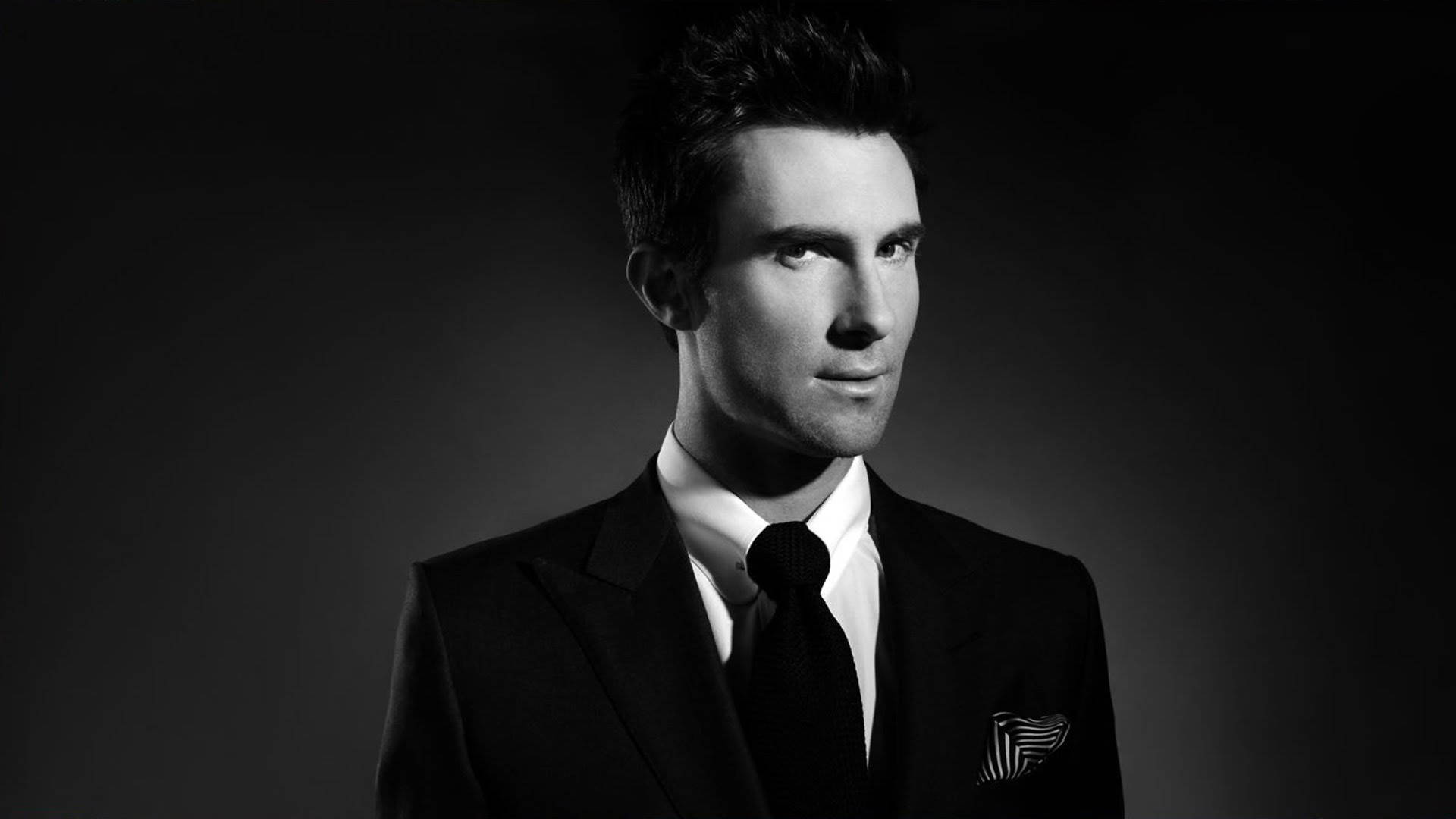 Adam Levine 桌布 壁紙 — 一位穿著黑色西裝和領帶、衣著講究的男士，在深色漸變背景前擺出姿勢，拍攝一張戲劇性的黑白肖像照。