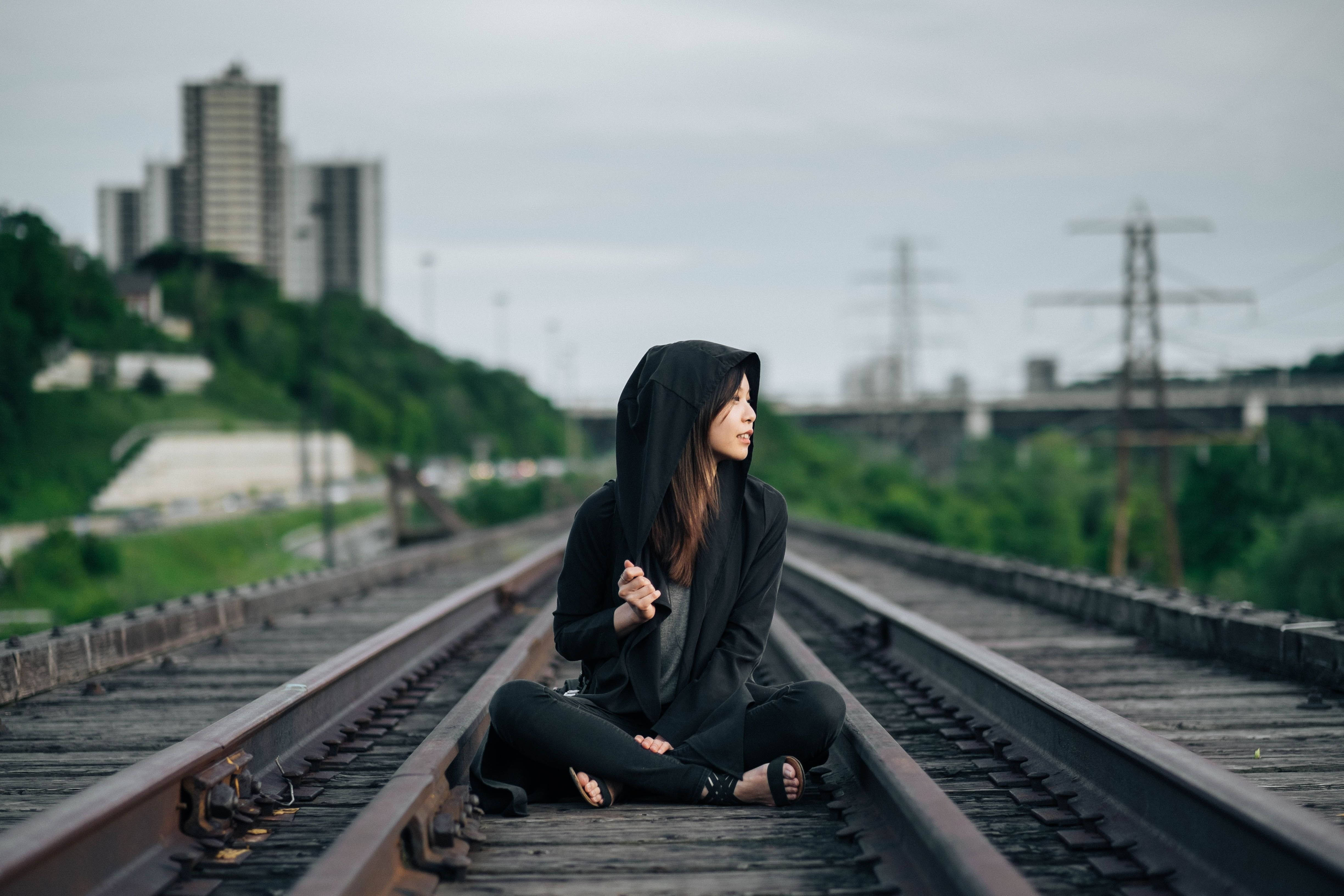 女孩 桌布 壁紙 — 一位穿著黑色連帽衫的年輕女性盤腿坐在磨損的火車軌道上，陰天的背景中有城市建築和綠色山丘。