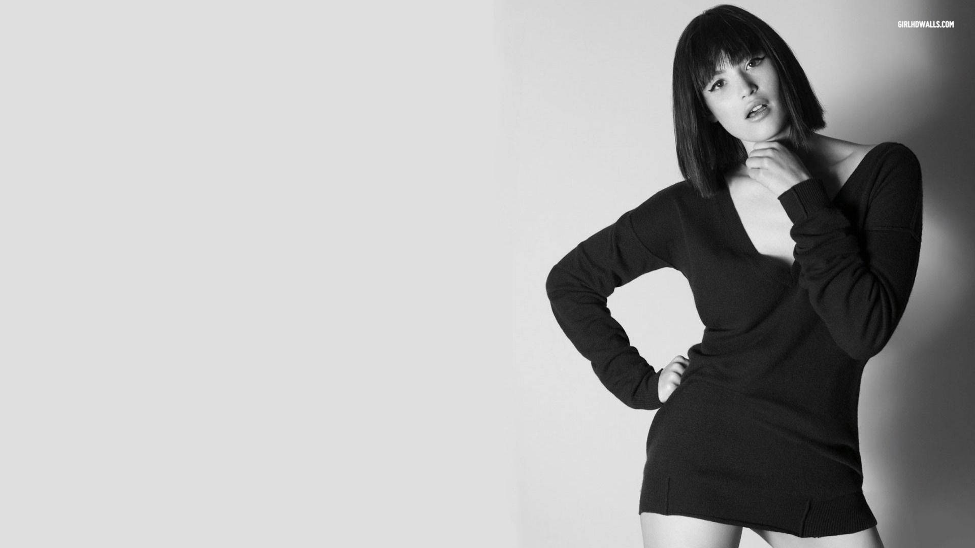 Gemma Arterton 桌布 壁紙 — 一位穿著黑色連衣裙的女性在簡約的黑白工作室拍攝中，自信地擺姿勢，背景是純灰色。