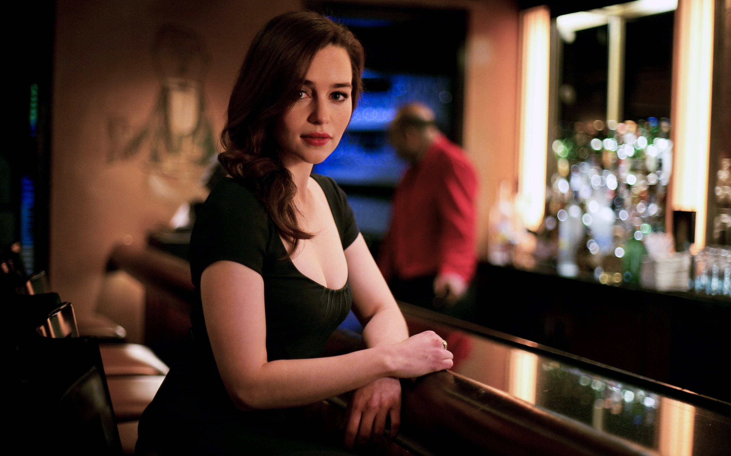 Emilia 桌布 壁紙 — 一位穿著黑色連衣裙的女性靠在光線昏暗的夜總會的吧檯上，背景有發光的瓶子和模糊的酒保。