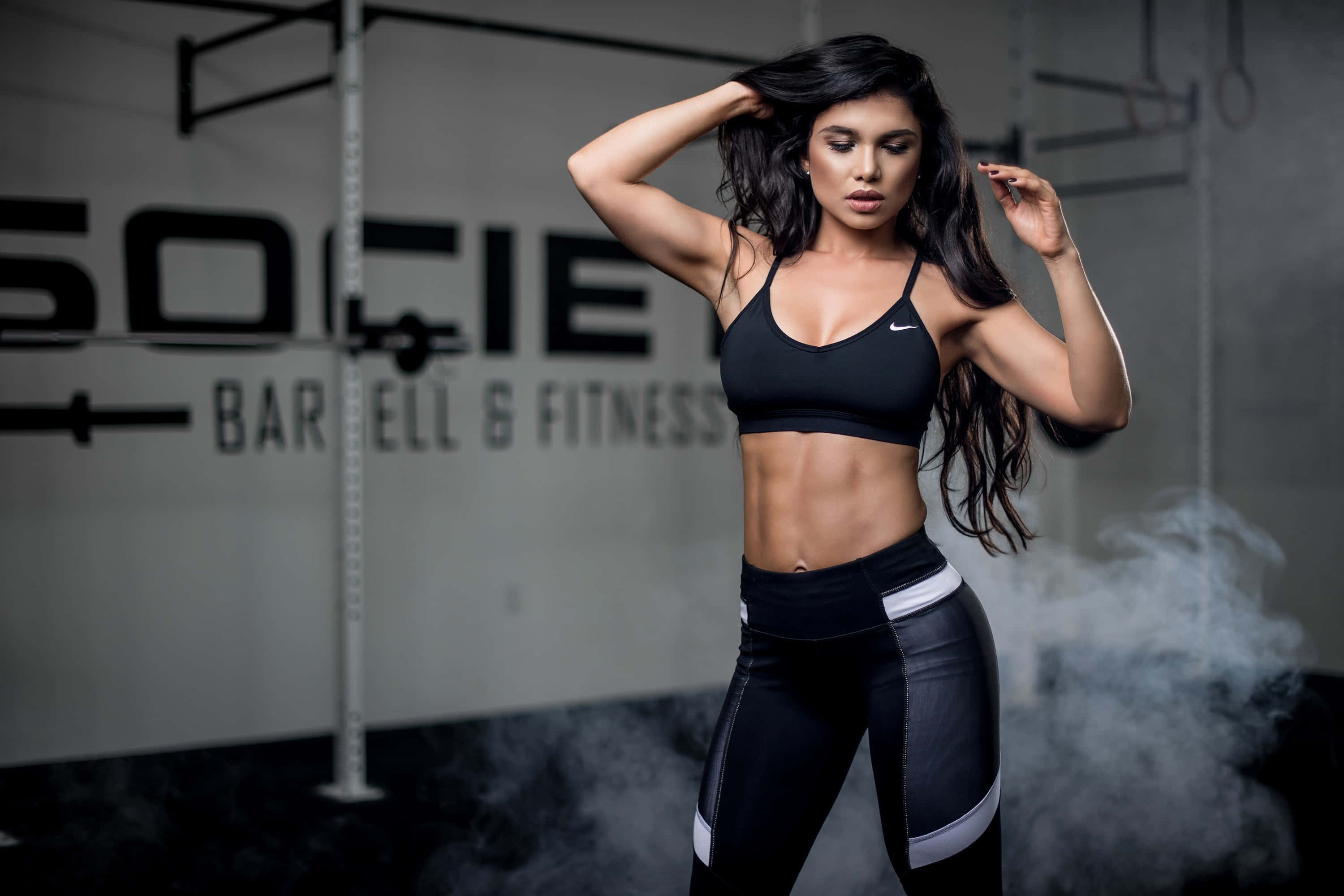 Fitness 桌布 壁紙 — 一位穿著黑色運動服的健美女性在煙霧瀰漫的健身房中自信地擺姿勢，牆上有'SOCIETY BARBELL & FITNESS'標誌。
