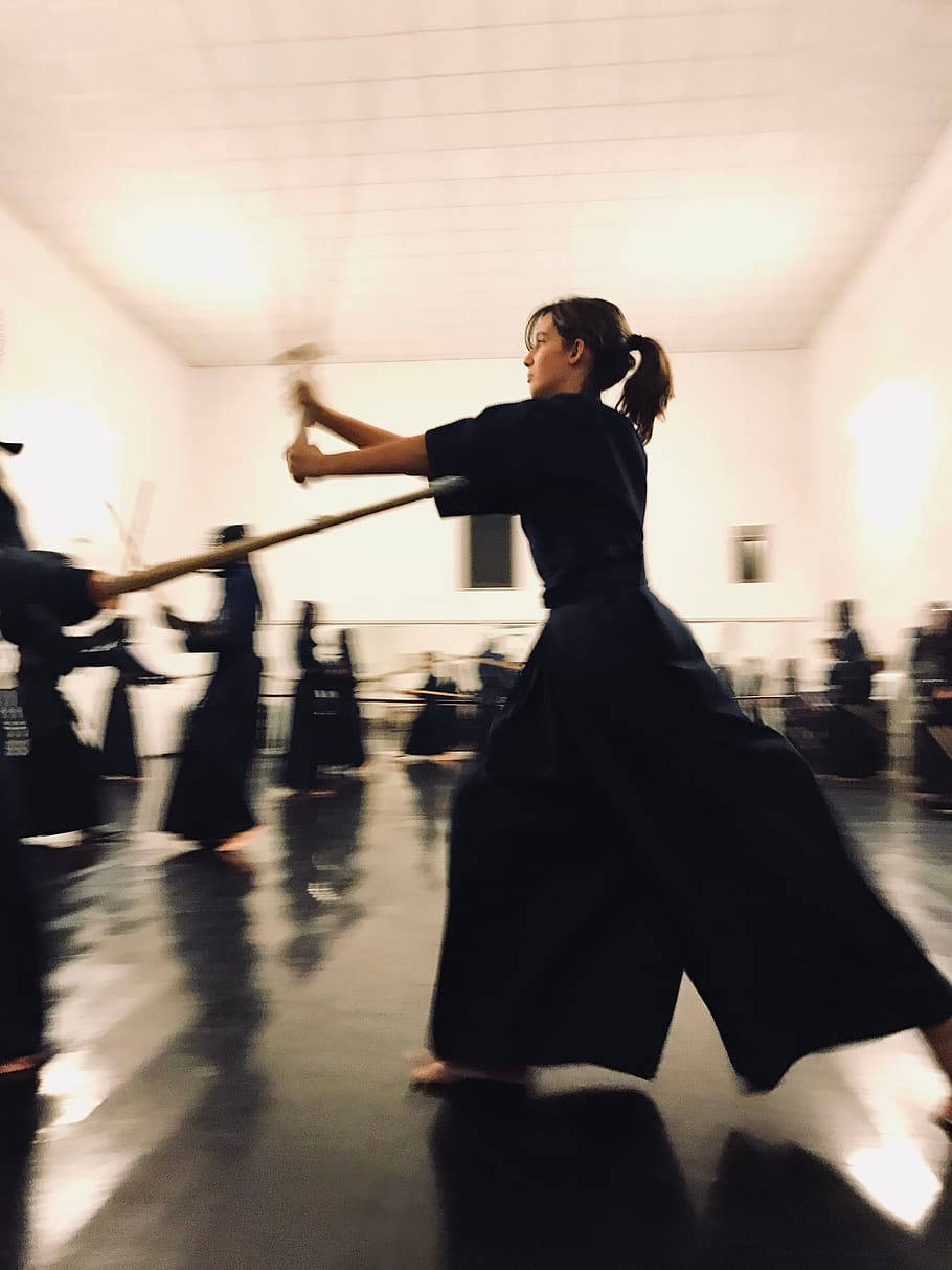 sword 桌布 壁紙 — 一位穿著黑色道服的武術家在光線充足、地板反光的道場中揮動木劍，帶有動態模糊效果。